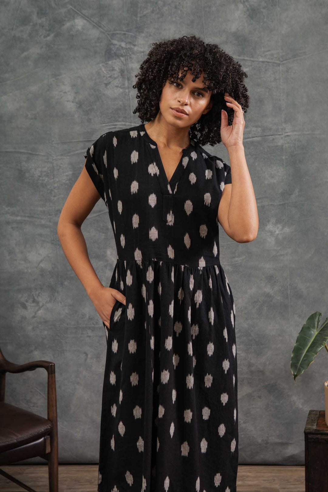 A woman with curly hair models the Bridge & Burn Ira Dress in Eclipse Ikat, a black pullover with white patterns and dolman sleeves. She stands indoors by a wooden chair and plant, hand in pocket, against a gray textured backdrop.
