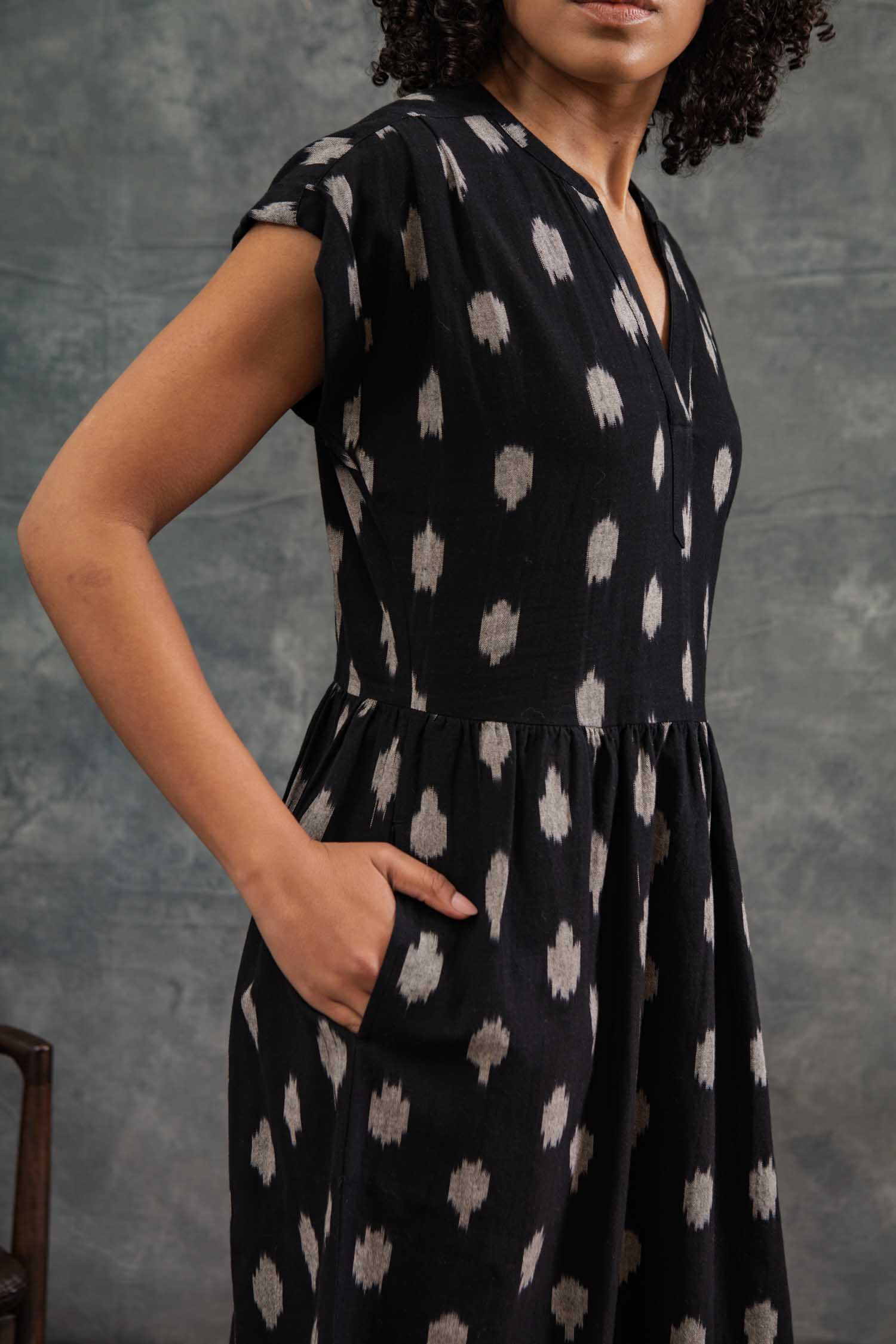 A woman stands with one hand in her pocket against a textured gray background, wearing the Bridge & Burn Ira Dress in Eclipse Ikat—a black 100% cotton pullover dress with short dolman sleeves and a light gray abstract pattern. Her face is partially out of frame.