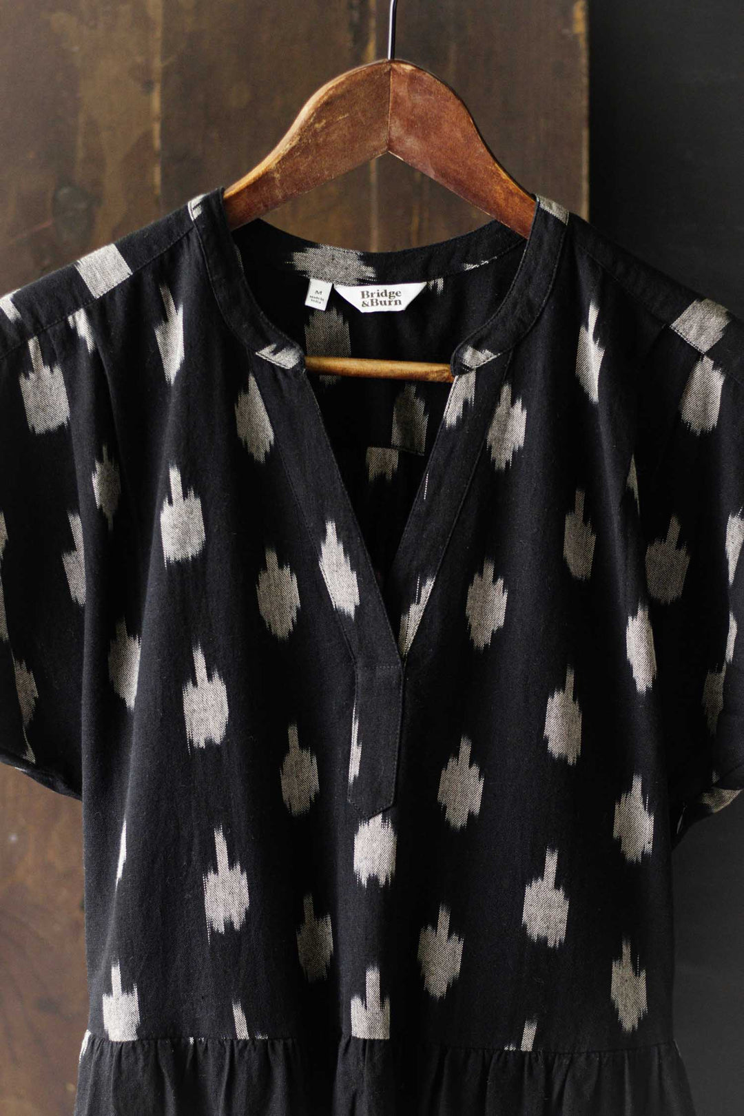 The Ira Dress / Eclipse Ikat by Bridge & Burn is a black 100% cotton pullover with short dolman sleeves, a notch neckline, and a white abstract dotted pattern, shown on a wooden hanger against a rustic brown backdrop.