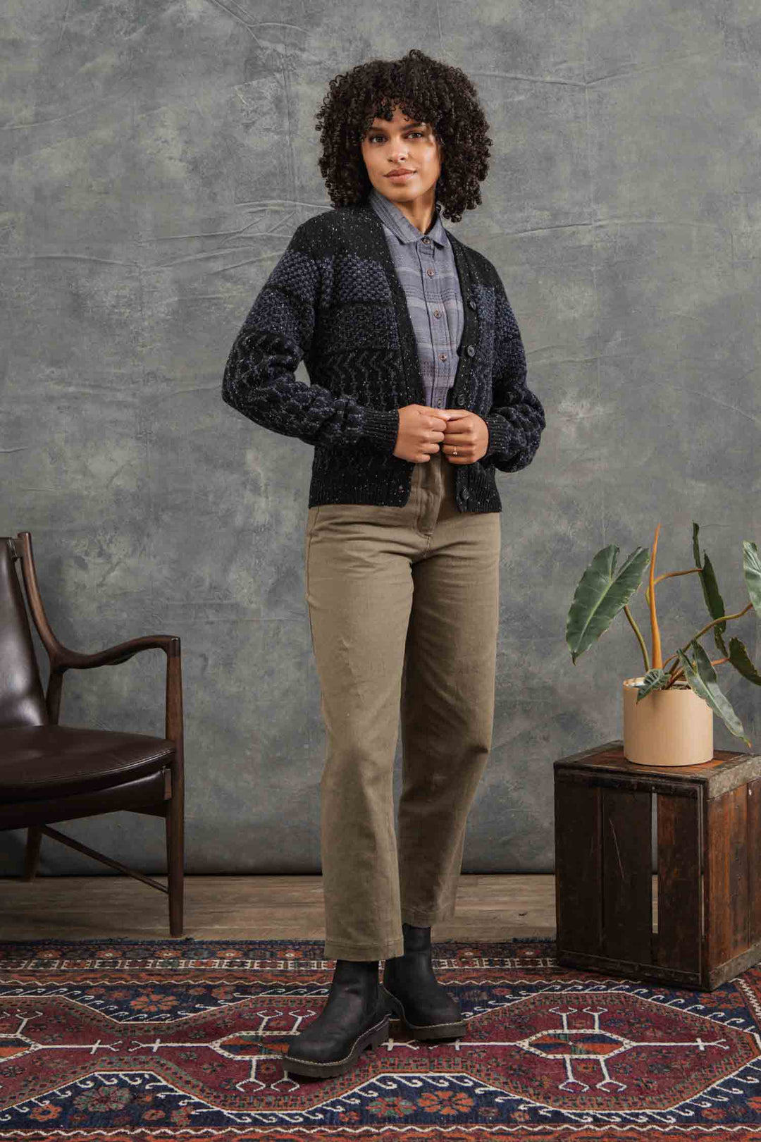 A woman with curly hair stands on a patterned rug indoors, wearing the Bridge & Burn Juneau Cardigan in Midnight Tweed over a gray plaid shirt, olive green pants, and black boots. A potted plant and chair are in the background by a gray wall.