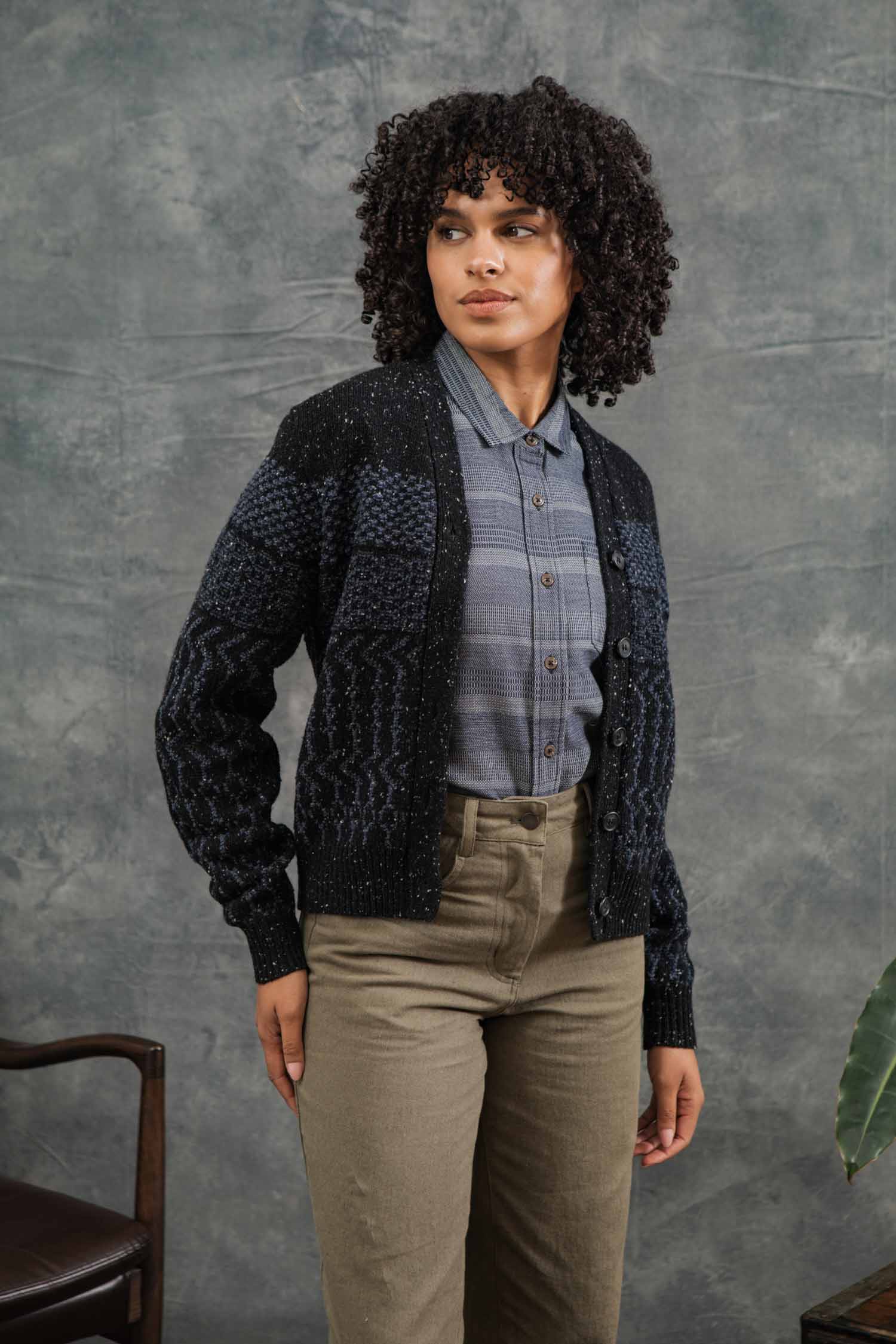 A woman with curly hair wears the Bridge & Burn Juneau Cardigan in Midnight Tweed over a gray plaid shirt and olive green pants, standing before a textured gray backdrop and gazing slightly to the side.