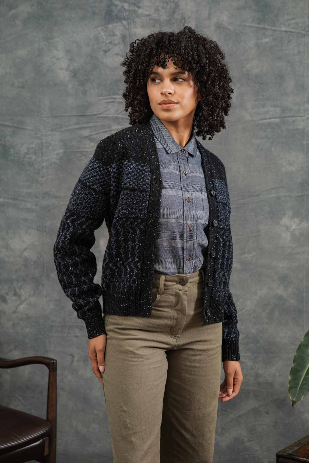 A woman with curly hair wears the Bridge & Burn Juneau Cardigan in Midnight Tweed over a gray plaid shirt and olive green pants, standing before a textured gray backdrop and gazing slightly to the side.
