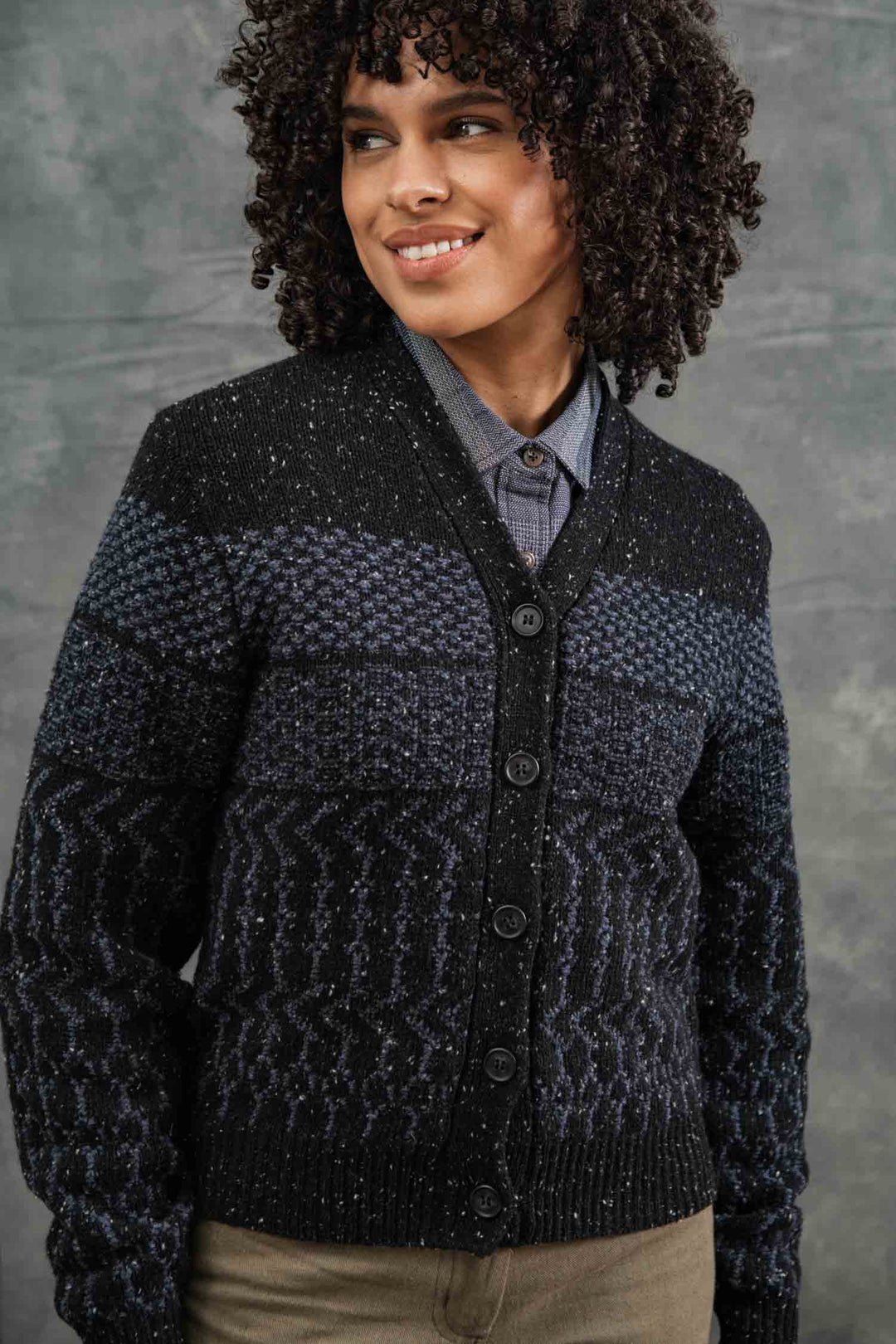 A person with curly hair smiles and looks to the side, wearing the Bridge & Burn Juneau Cardigan in Midnight Tweed over a collared shirt, standing in front of a gray background.