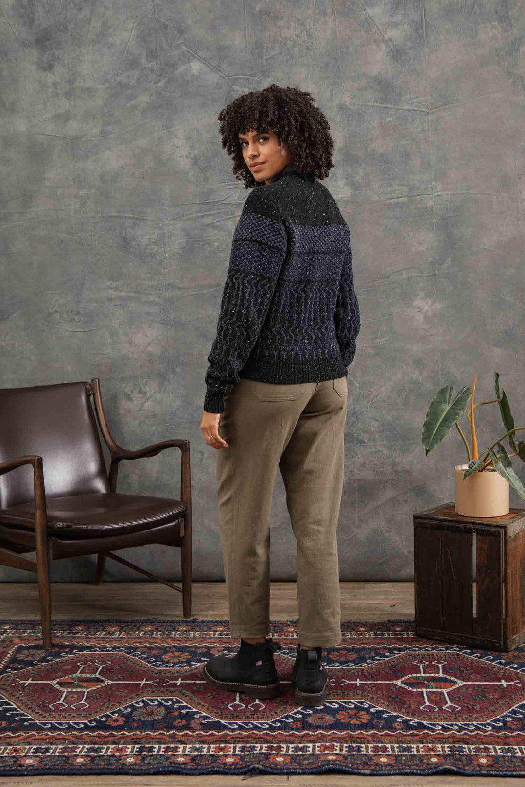 A person with curly hair wears the Bridge & Burn Juneau Cardigan in Midnight Tweed and khaki pants, standing in a room with a patterned rug, wooden chair, and small plant on a wooden box, looking back over their shoulder.