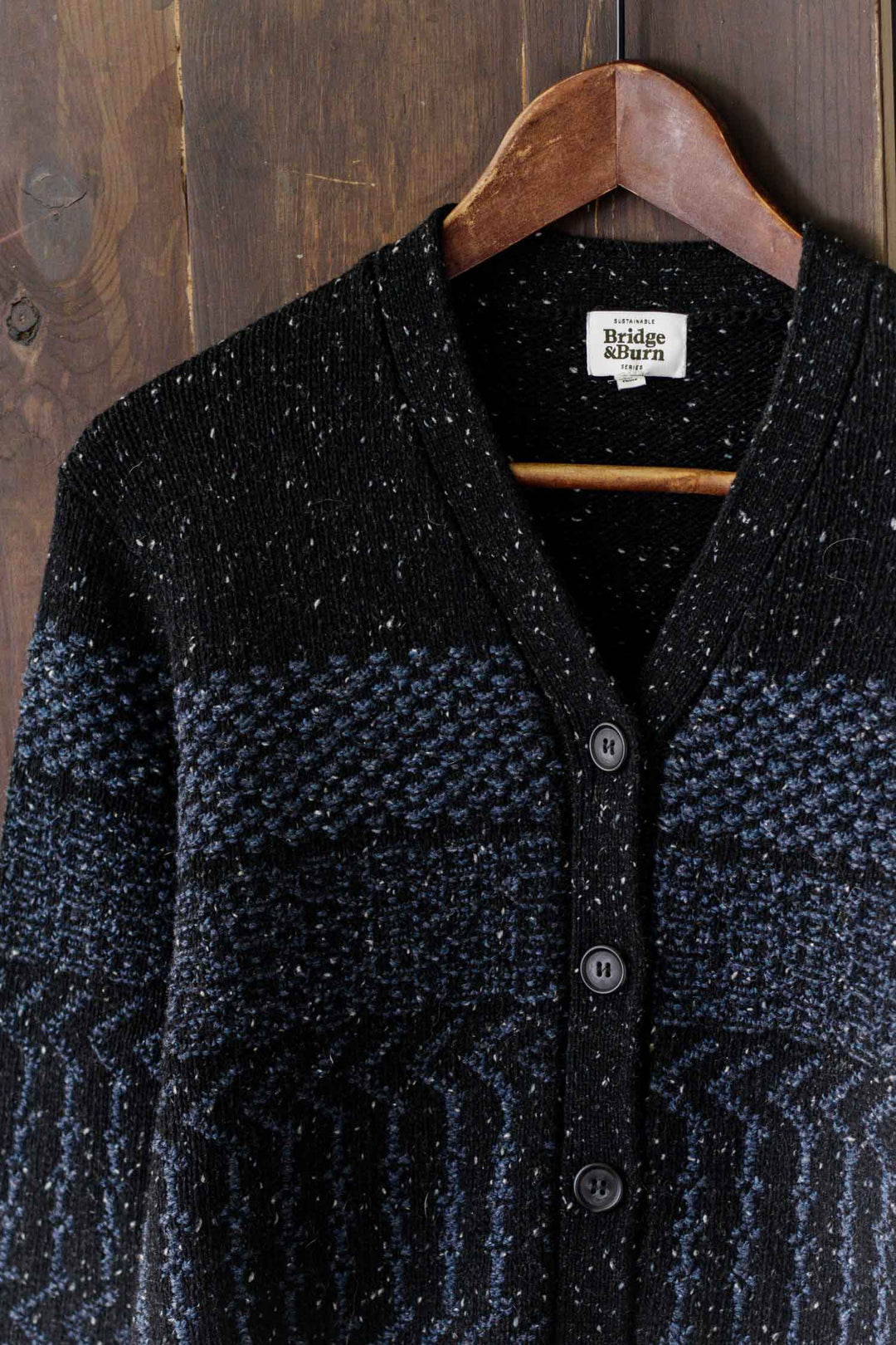 The Bridge & Burn Juneau Cardigan in Midnight Tweed, featuring subtle blue and gray stripes with black buttons, hangs on a wooden hanger against a rustic wood background.