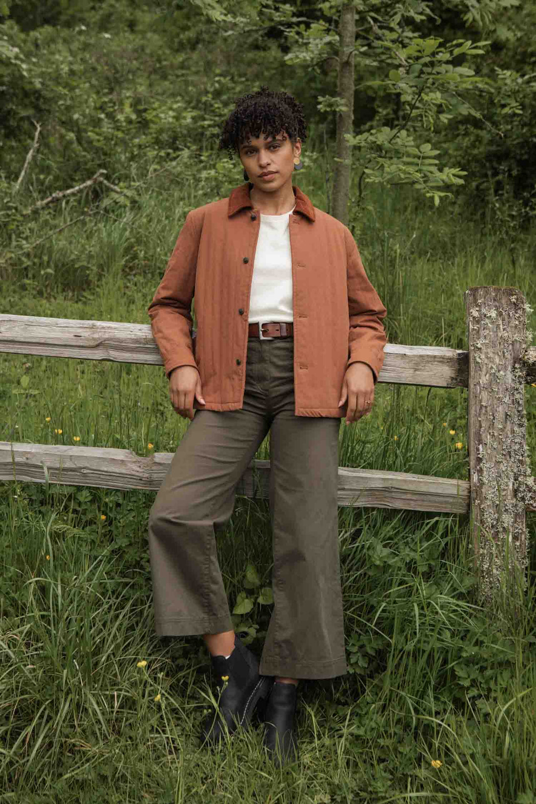 A person stands against a wooden fence in a grassy area, wearing the Bridge & Burn Kenton Jacket in Sienna, paired with a white t-shirt, olive green wide-leg pants, and black boots, with greenery and trees in the background.
