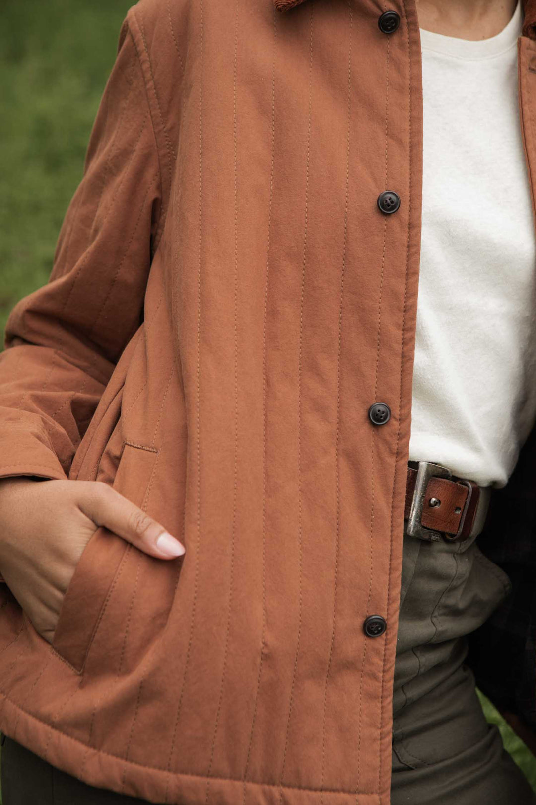 Wearing Bridge & Burn's Kenton Jacket in Sienna—a rust-colored quilted jacket with a corduroy collar—over a white shirt and olive trousers with a brown belt, the person stands outdoors against a softly blurred grassy background.