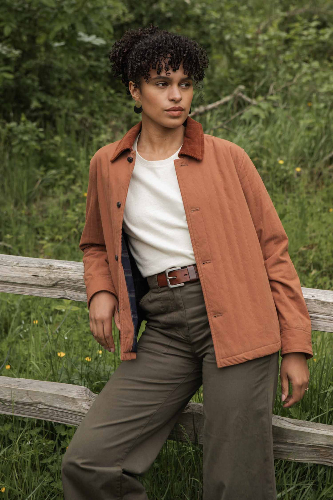 A woman with curly hair stands by a wooden fence, wearing the Bridge & Burn Kenton Jacket in Sienna over a white shirt with olive green pants and a brown belt, surrounded by greenery and grass.