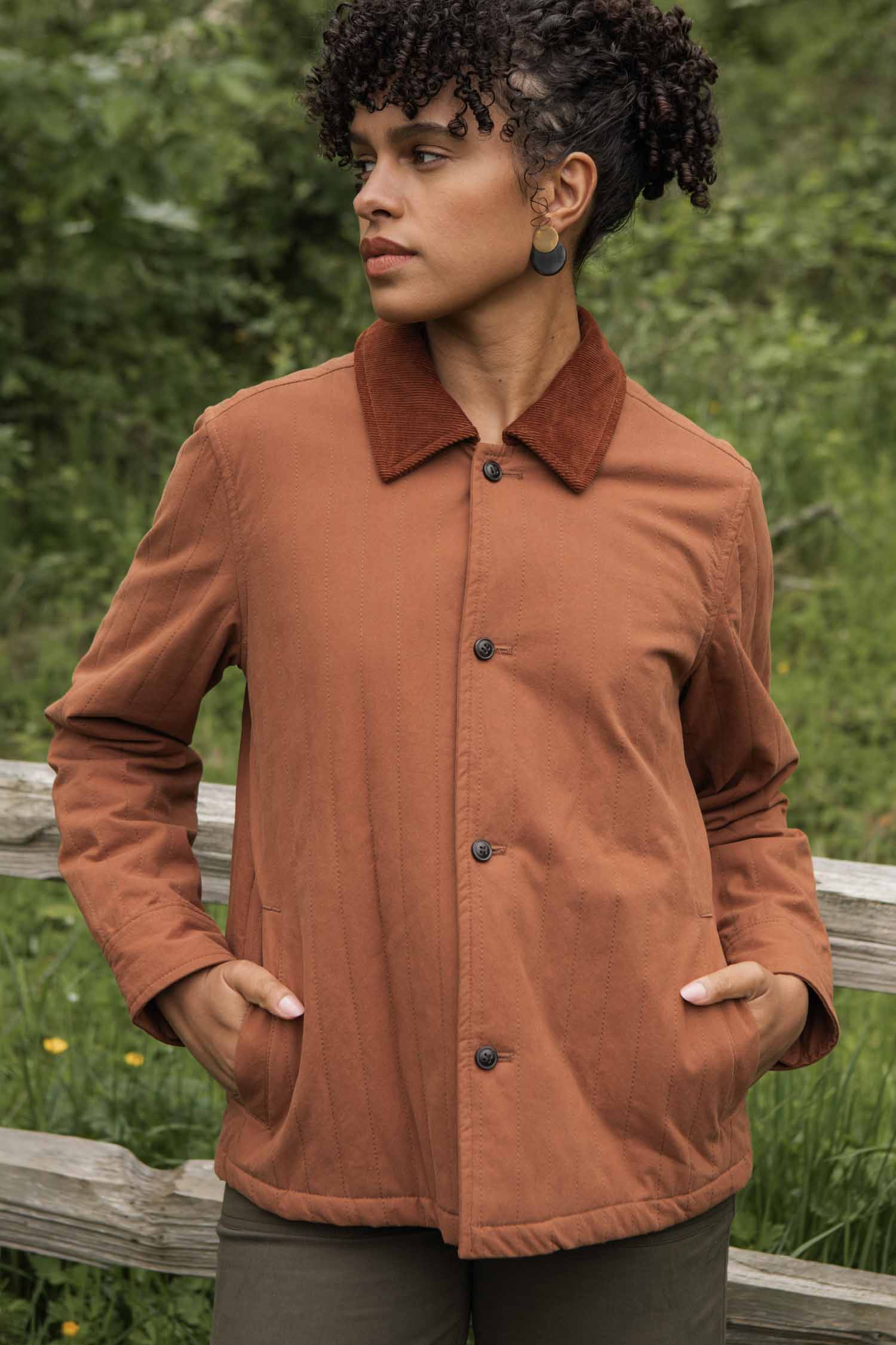 A person with short curly hair stands outdoors in front of greenery and a wooden fence, wearing the Bridge & Burn Kenton Jacket in Sienna with a corduroy collar and dark pants, hands in pockets, looking to the side.