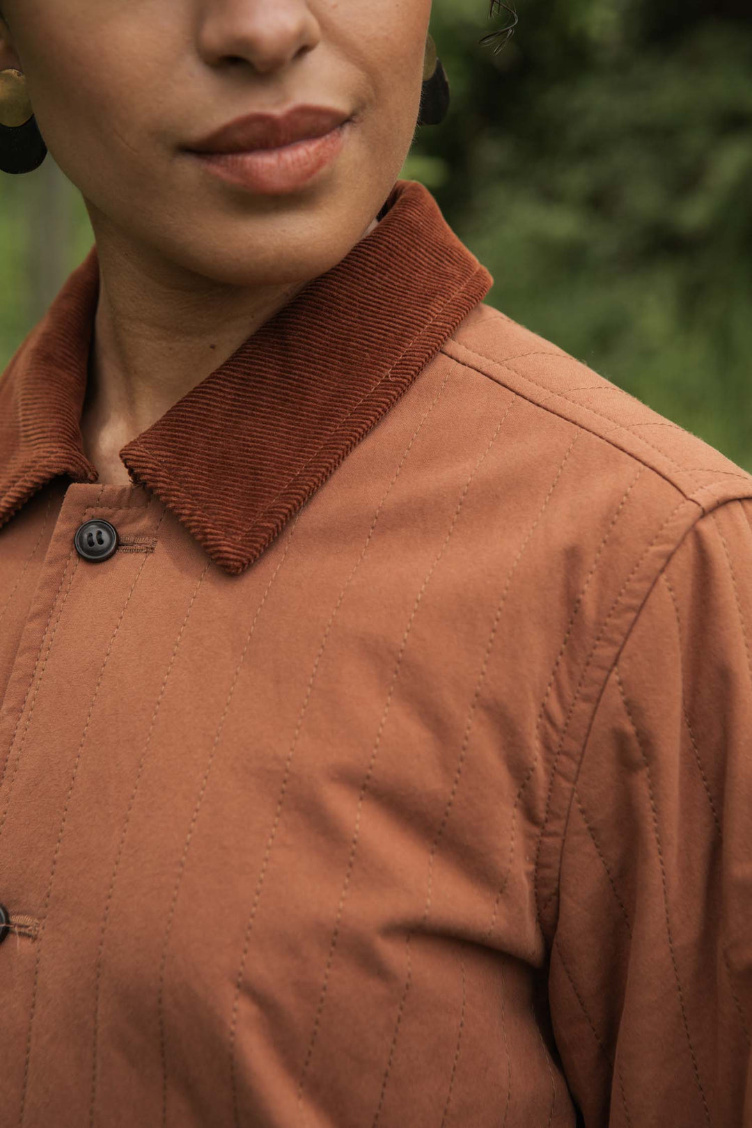 A close-up of a person wearing the Bridge & Burn Kenton Jacket in Sienna, featuring a corduroy collar and detailed stitching outdoors. Only the lower half of the face and upper torso are visible against a blurred green background.
