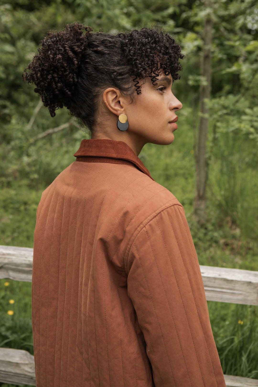 A person with curly hair in a puff, wearing the Bridge & Burn Kenton Jacket in Sienna and oval earrings, stands outdoors before a wooden fence and greenery, gazing to the right.