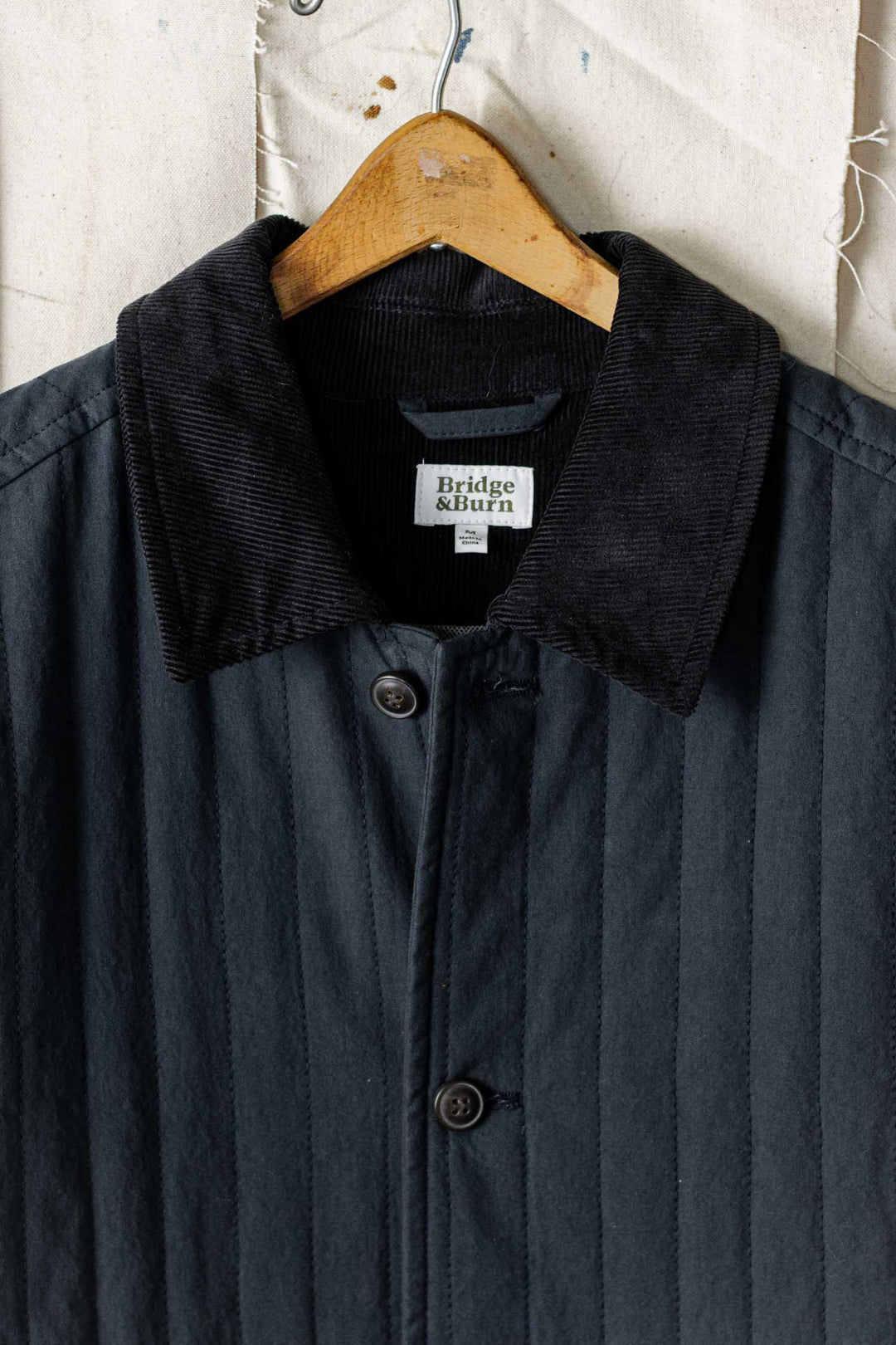 A dark, quilted Bridge & Burn Kenton Jacket in Slate with a black corduroy collar hangs on a wooden hanger against a light, textured fabric backdrop, highlighting this lightweight jacket’s timeless style.