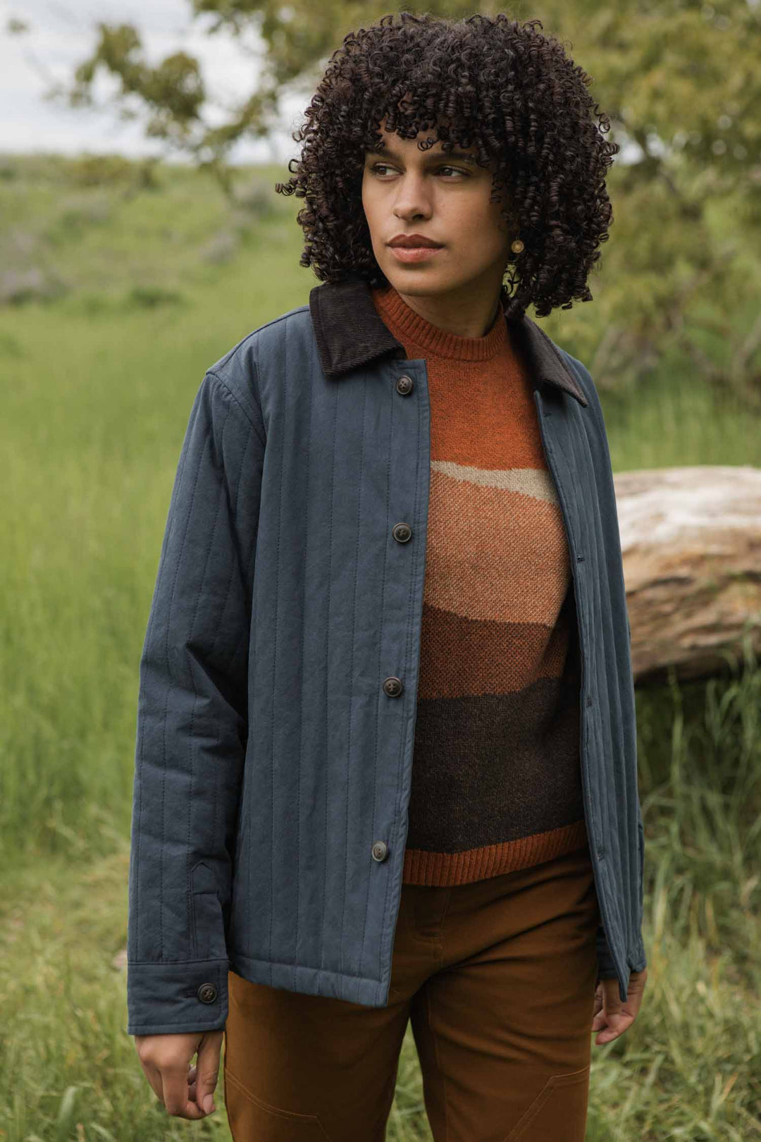A person with curly hair stands outdoors in a grassy area, wearing the Bridge & Burn Kenton Jacket in Slate over a brown and orange striped sweater and brown pants. A fallen tree and greenery are visible in the background.