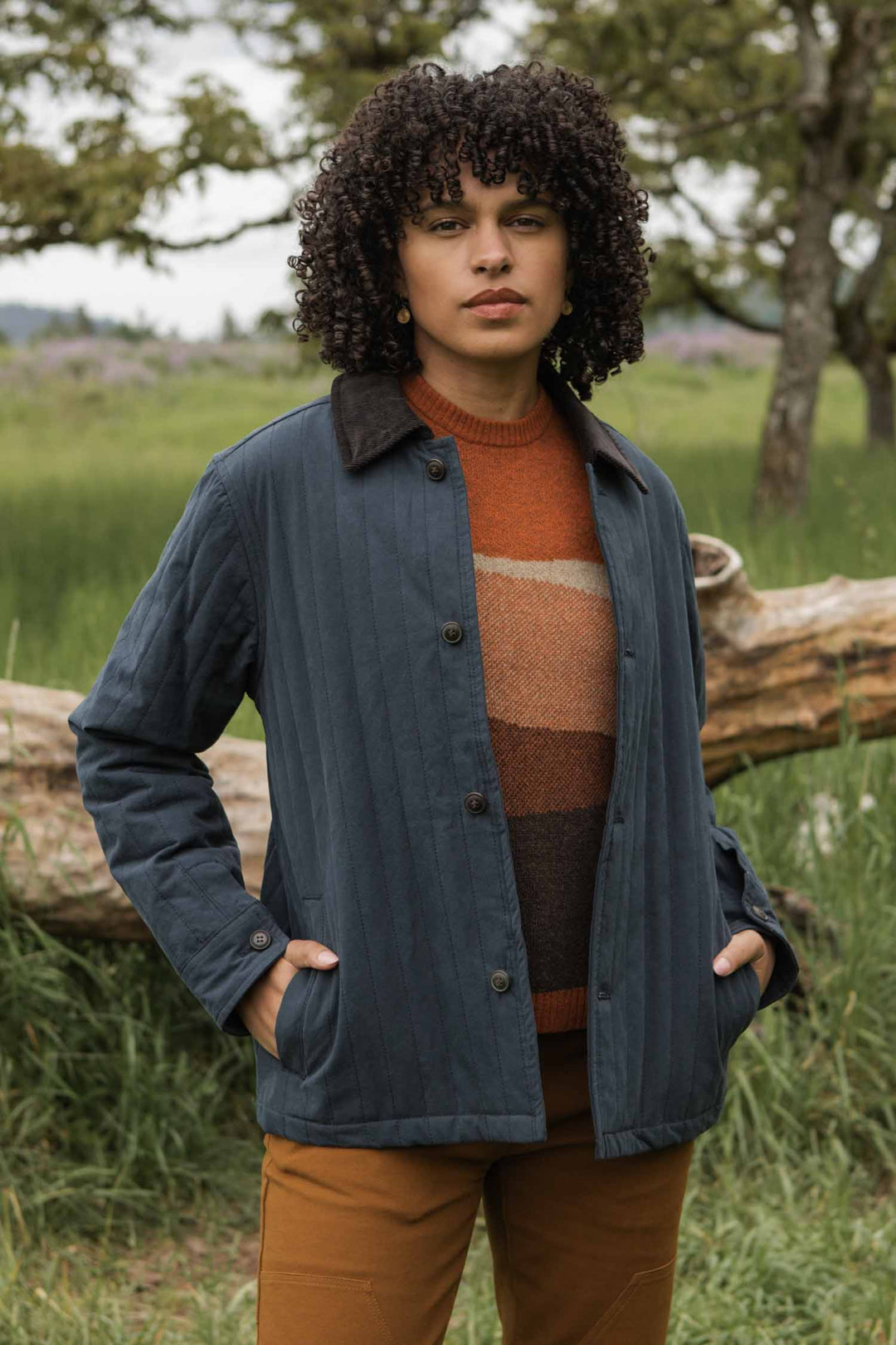 A person with curly hair stands outdoors amid greenery, hands in pockets, wearing brown pants, a striped sweater, and the Bridge & Burn Kenton Jacket in Slate—a blue quilted jacket featuring a corduroy collar.