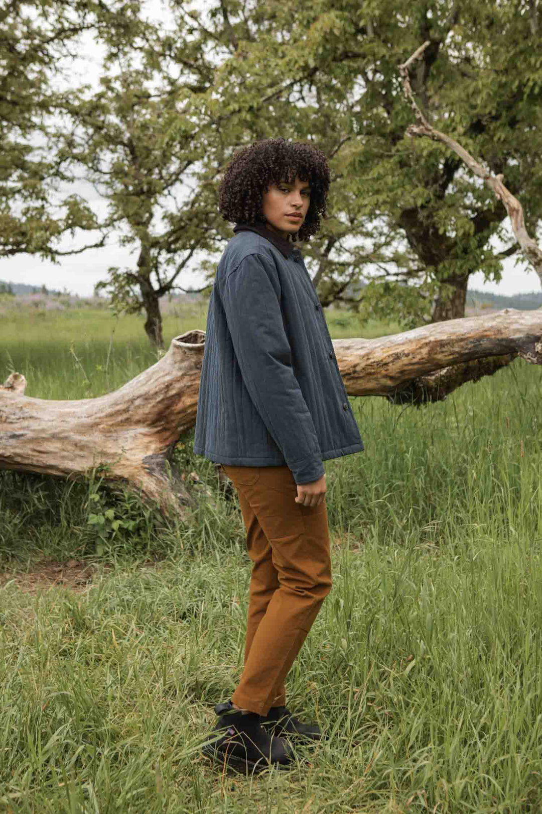 Outdoors on grass by a fallen tree, a person with curly hair wears the Bridge & Burn Kenton Jacket in Slate, paired with brown pants and black shoes; green trees and meadow form the background.