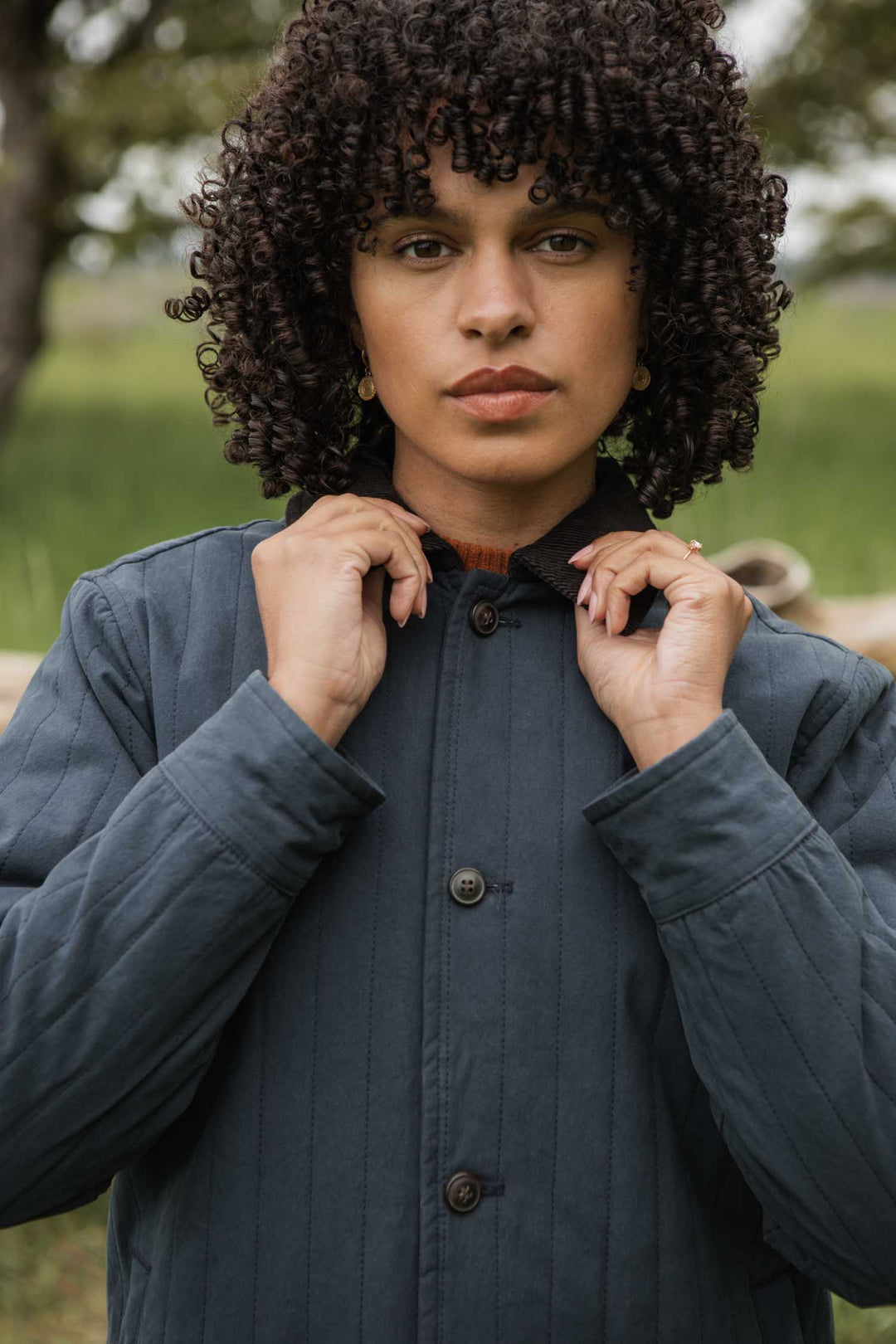 Outdoors, a person with curly hair adjusts the corduroy collar of the Bridge & Burn Kenton Jacket in Slate while facing the camera. Blurred green grass and trees fill the background.