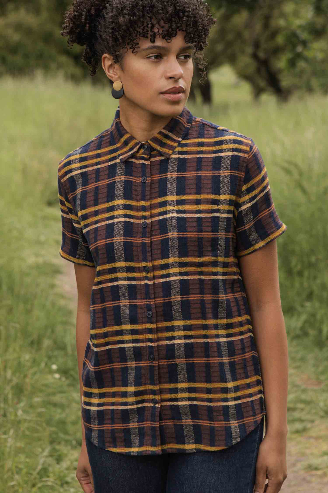 A person stands on a grassy path wearing the Bridge & Burn Lana Shirt in Desert Dawn Plaid, a relaxed fit, short sleeve button-up made from organic cotton, paired with dark jeans. They have curly hair and look to the side.