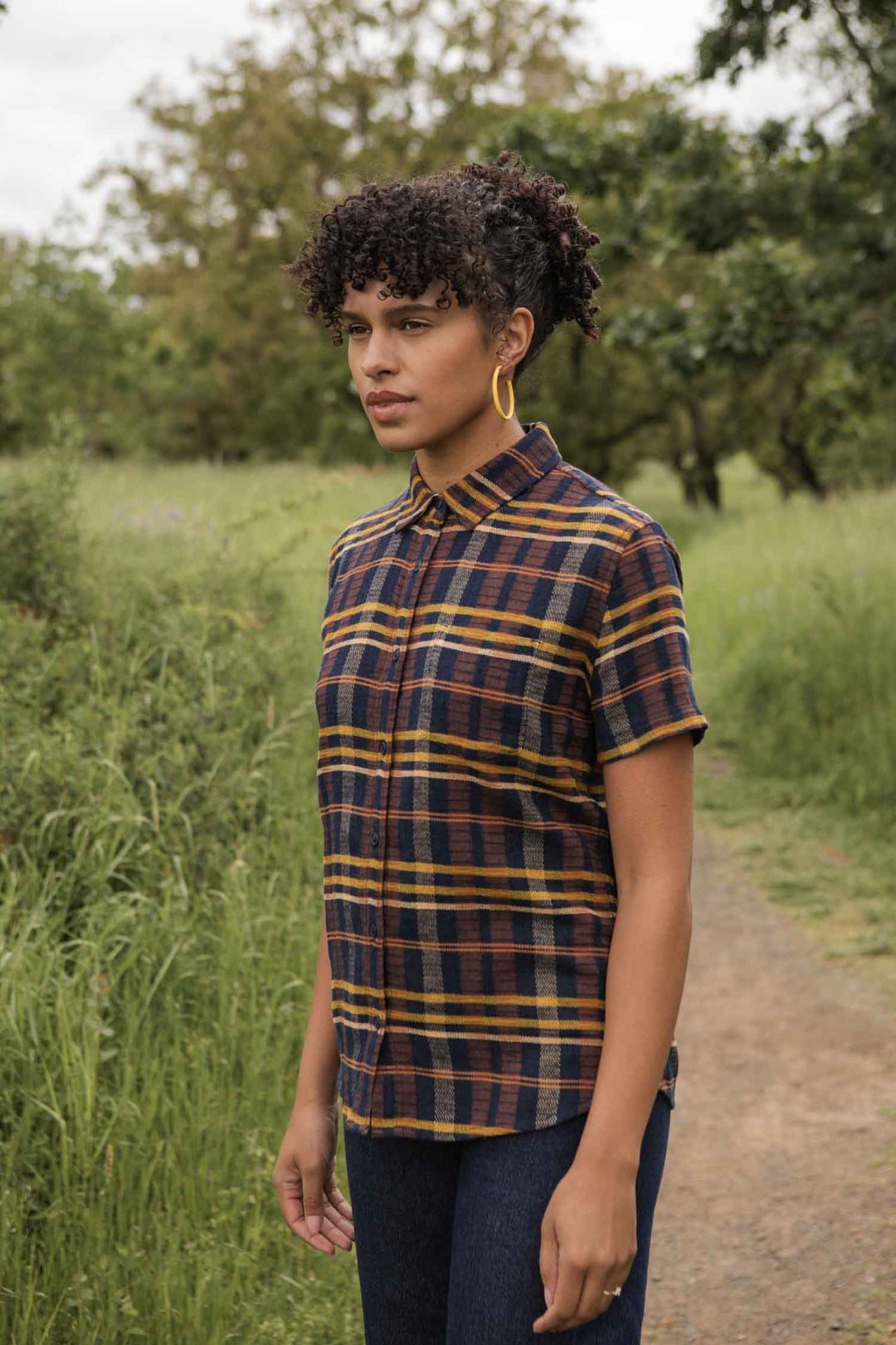 A person with short curly hair, hoop earrings, and a relaxed fit Bridge & Burn Lana Shirt in Desert Dawn Plaid stands on a dirt path surrounded by greenery, looking slightly to the side.