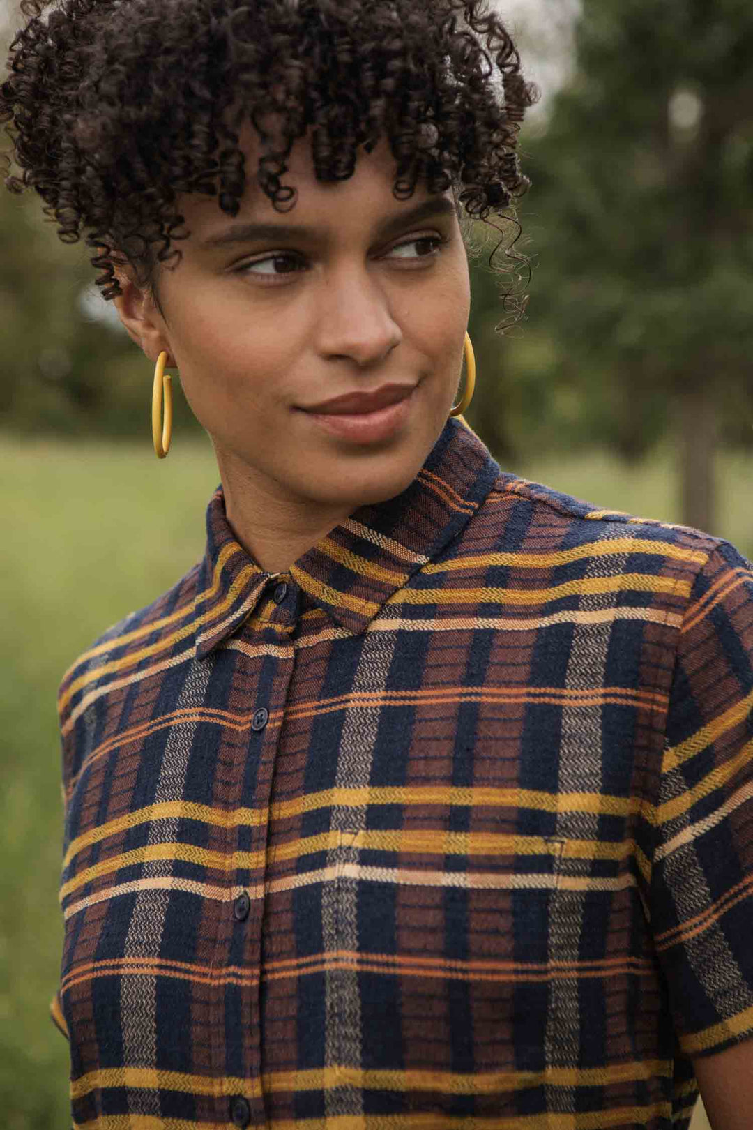 A person with short curly hair and gold hoop earrings wears the Bridge & Burn Lana Shirt in Desert Dawn Plaid, featuring blue, brown, and yellow stripes, while standing outdoors amidst greenery and trees.
