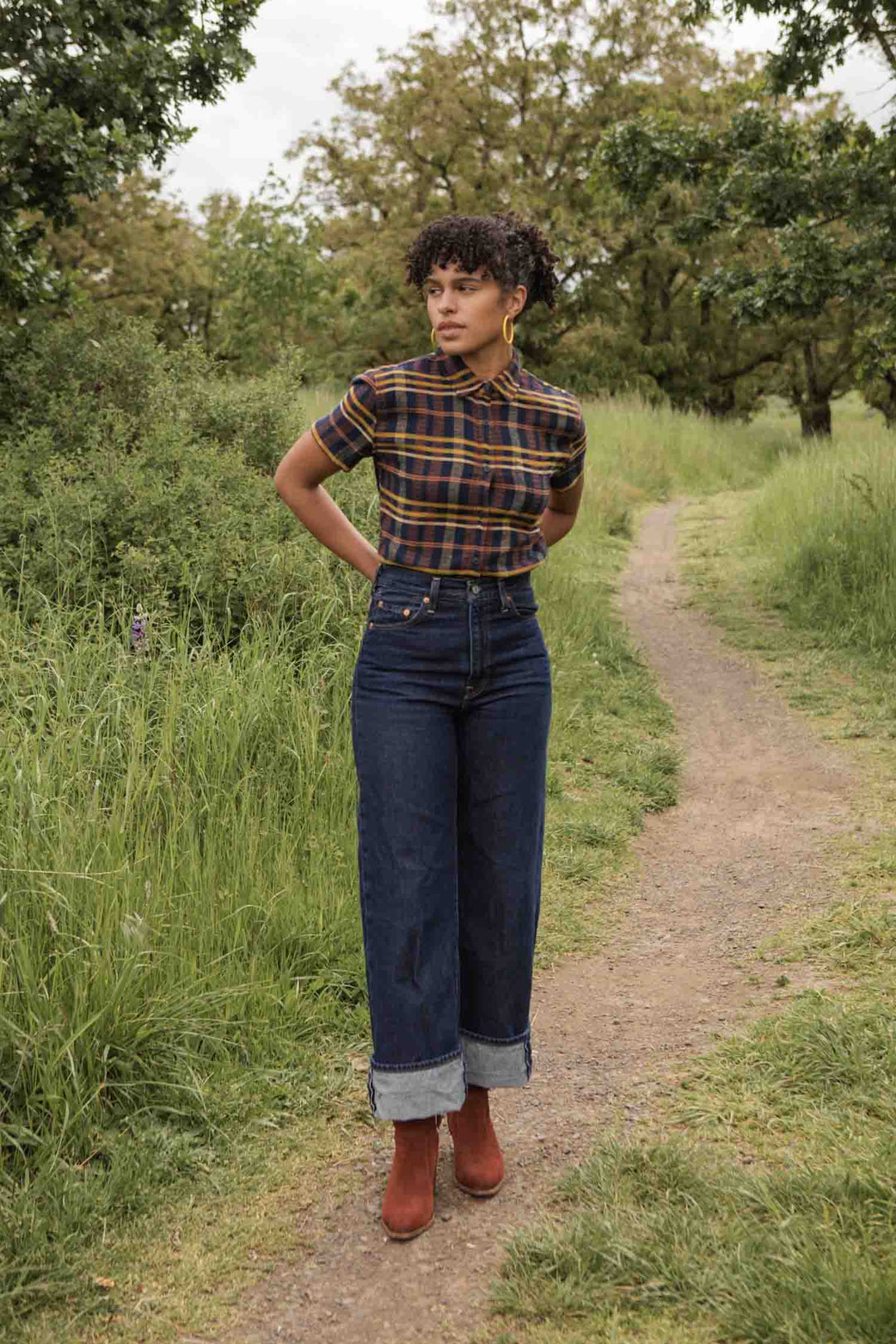 A person stands on a dirt path surrounded by greenery, wearing the Bridge & Burn Lana Shirt in Desert Dawn Plaid, high-waisted cuffed jeans, and brown boots. Their hands are behind their back as they look slightly to the side.