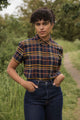 A person stands outdoors on a path surrounded by greenery, wearing the Bridge & Burn Lana Shirt in Desert Dawn Plaid, paired with high-waisted jeans and gold hoop earrings. They have short curly hair and a calm expression.