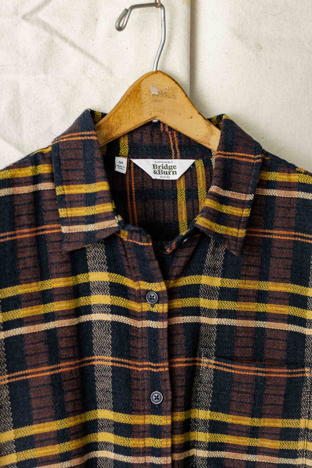 The Bridge & Burn Lana Shirt in Desert Dawn Plaid, a relaxed fit short sleeve button-up with a collar and front buttons in brown, yellow, and navy plaid, hangs on a wooden hanger against a light fabric background.