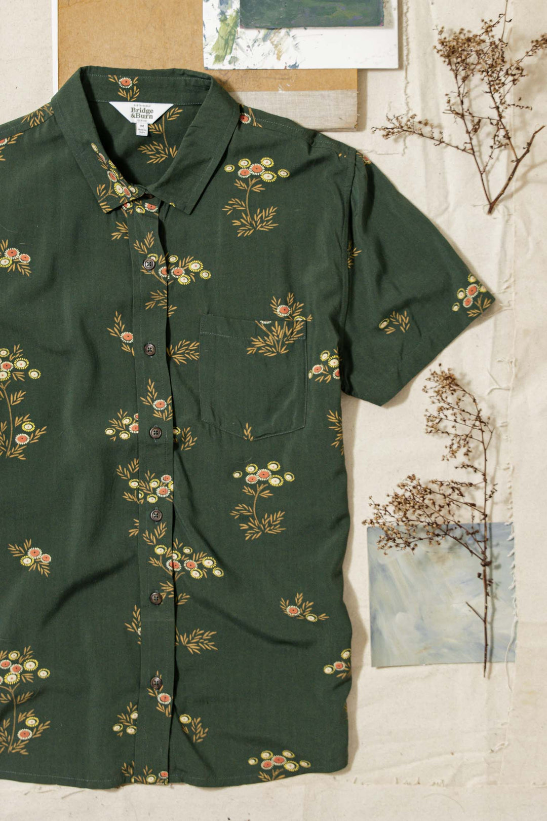 The Bridge & Burn Lana Shirt in Woodland Bloom—a dark green relaxed fit short sleeve button up with a yellow and orange floral print—rests on a beige surface with dried twigs and art. Made from an eco-friendly cotton alternative.