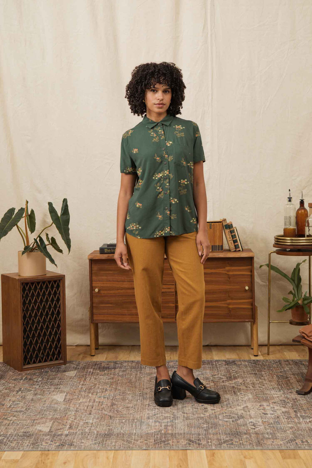 A woman stands indoors on a rug, wearing the Bridge & Burn Lana Shirt in Woodland Bloom, paired with mustard yellow pants and black loafers. Behind her is a mid-century wooden cabinet decorated with books, plants, and decor.