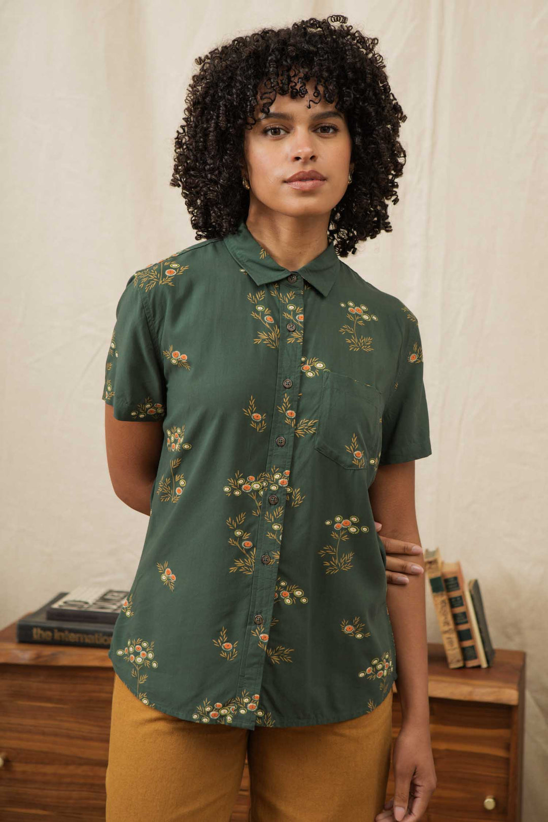 A person with curly hair wears the Bridge & Burn Lana Shirt in Woodland Bloom, a green, short-sleeve floral button-up in a relaxed fit, paired with mustard-colored pants and standing indoors by a beige backdrop and wooden cabinet with books.