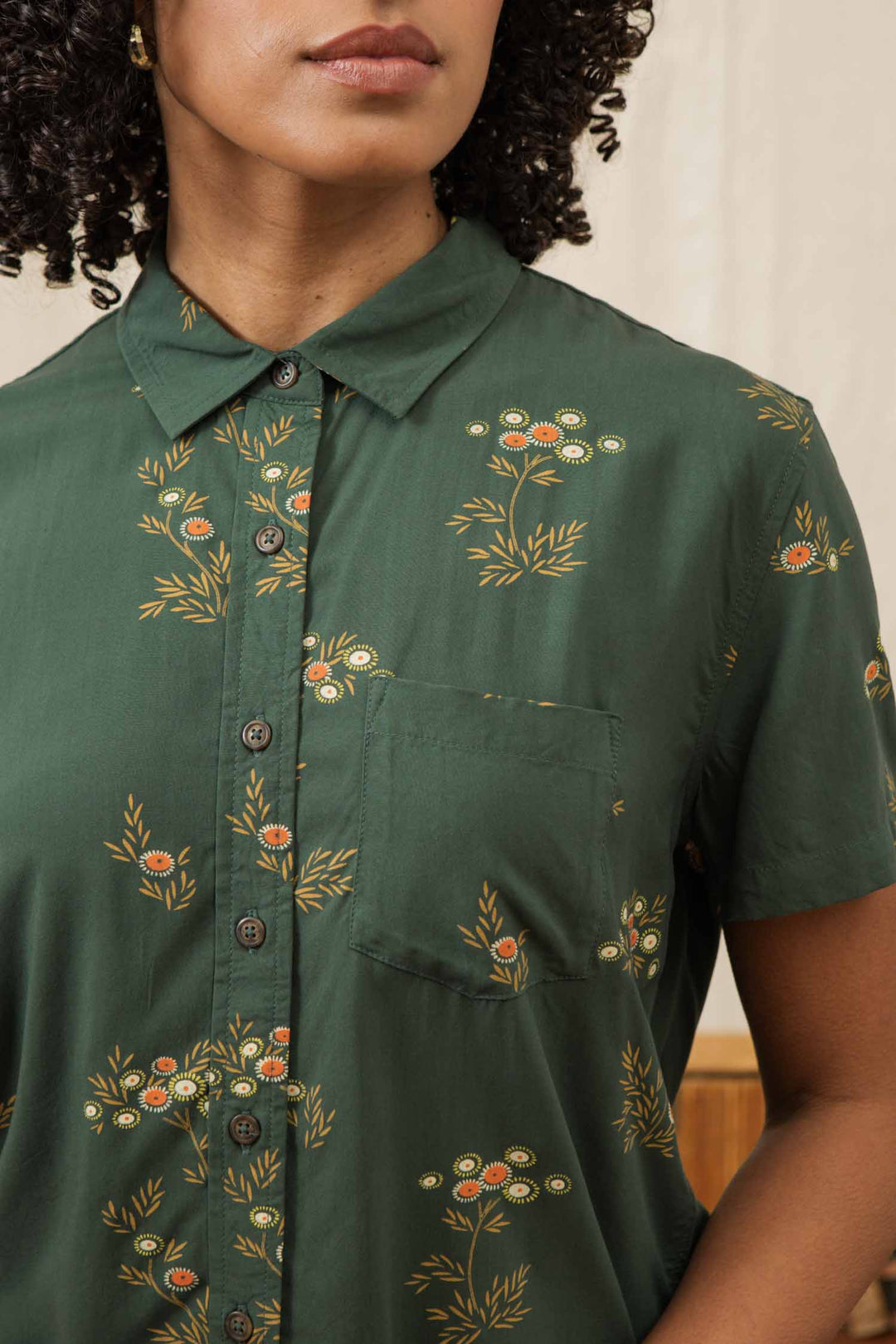 A woman wears the Bridge & Burn Lana Shirt in Woodland Bloom, a relaxed fit, dark green short sleeve button-up with a small pocket and white and orange floral print. Only her lower face and upper torso are visible.