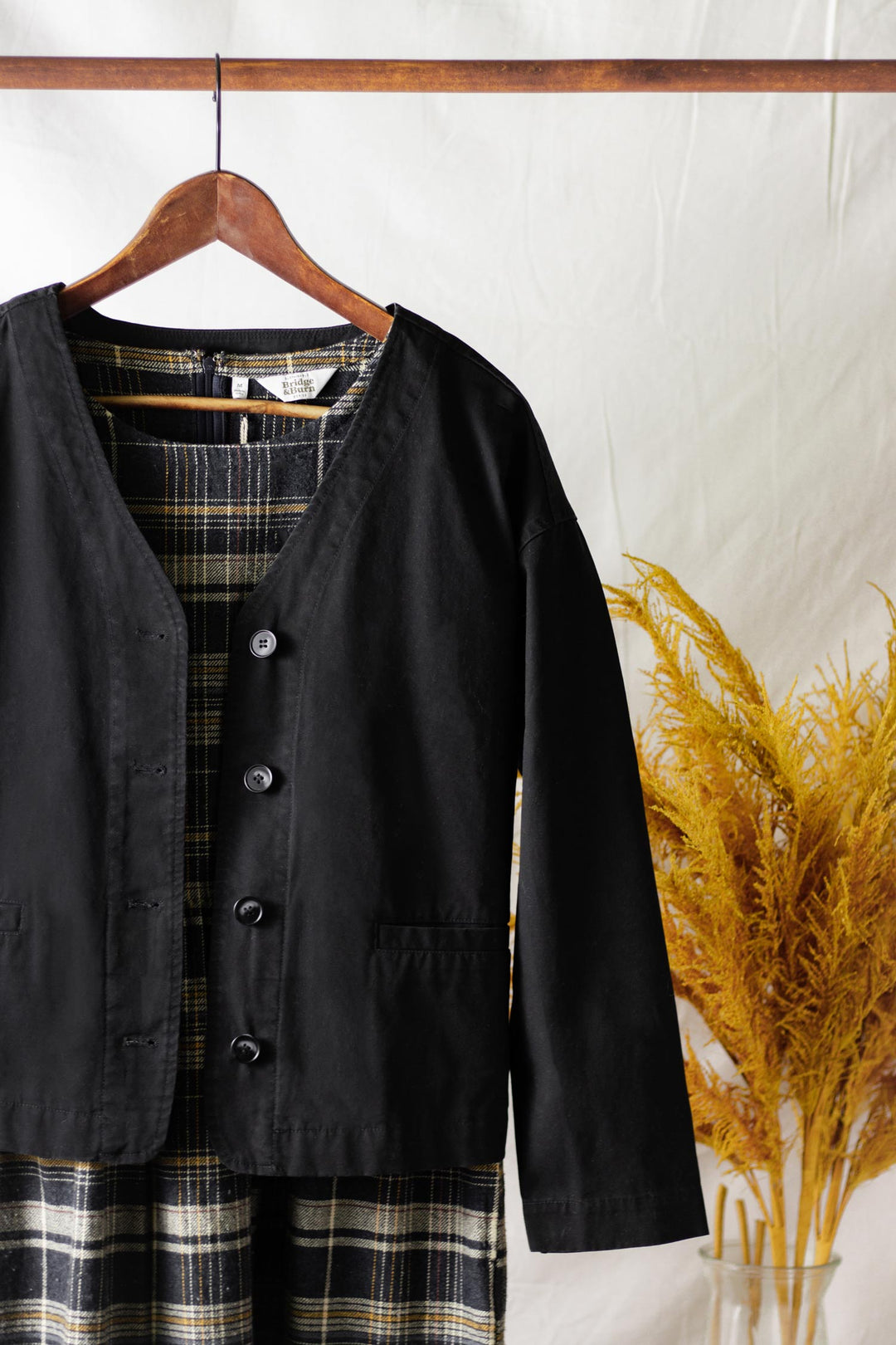 The Bridge & Burn Lee Jacket in black canvas, featuring a modern boxy fit, hangs on a wooden hanger over a plaid dress against a white fabric background. A vase with dried yellow plants sits to the right of the display.