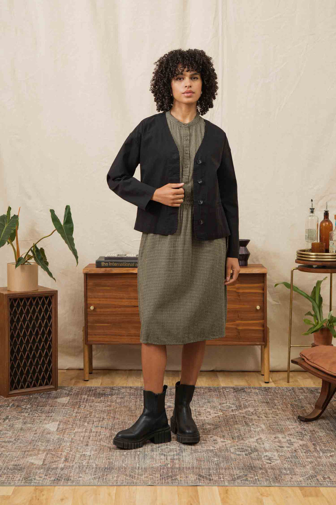 A woman with curly hair stands indoors on a rug, wearing a green plaid dress, black boots, and the Bridge & Burn Lee Jacket in Black Canvas. She is in front of a mid-century console table adorned with plants and bottles.