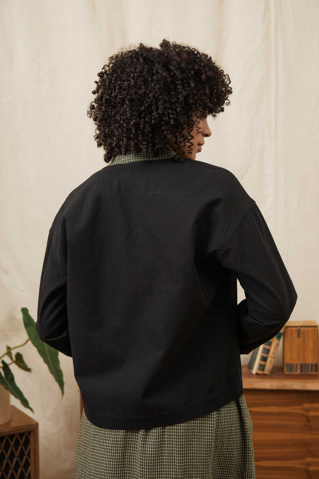 A person with curly hair wears the Bridge & Burn Lee Jacket in Black Canvas over a green checkered shirt, standing with their back to the camera in a room with neutral decor and a vintage radio on a wooden table.