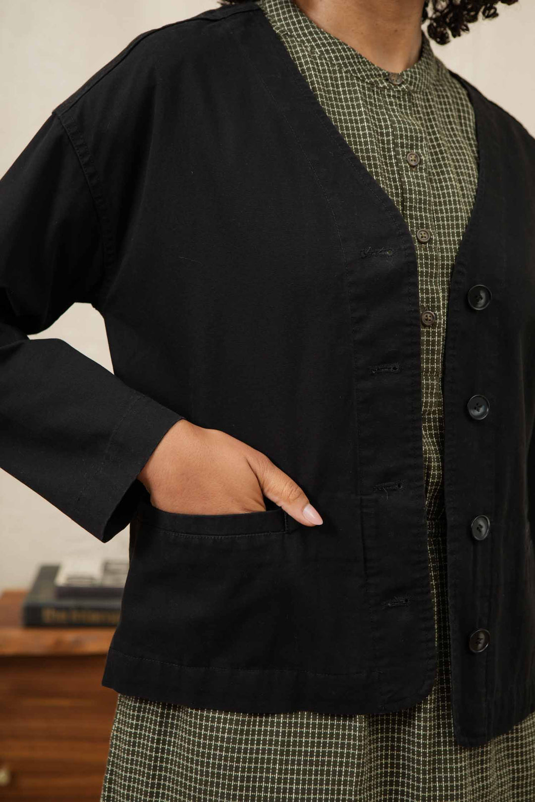 A person wears the Bridge & Burn Lee Jacket in black canvas over a green checkered dress, standing with one hand in a pocket. Only the torso and part of an arm are visible.