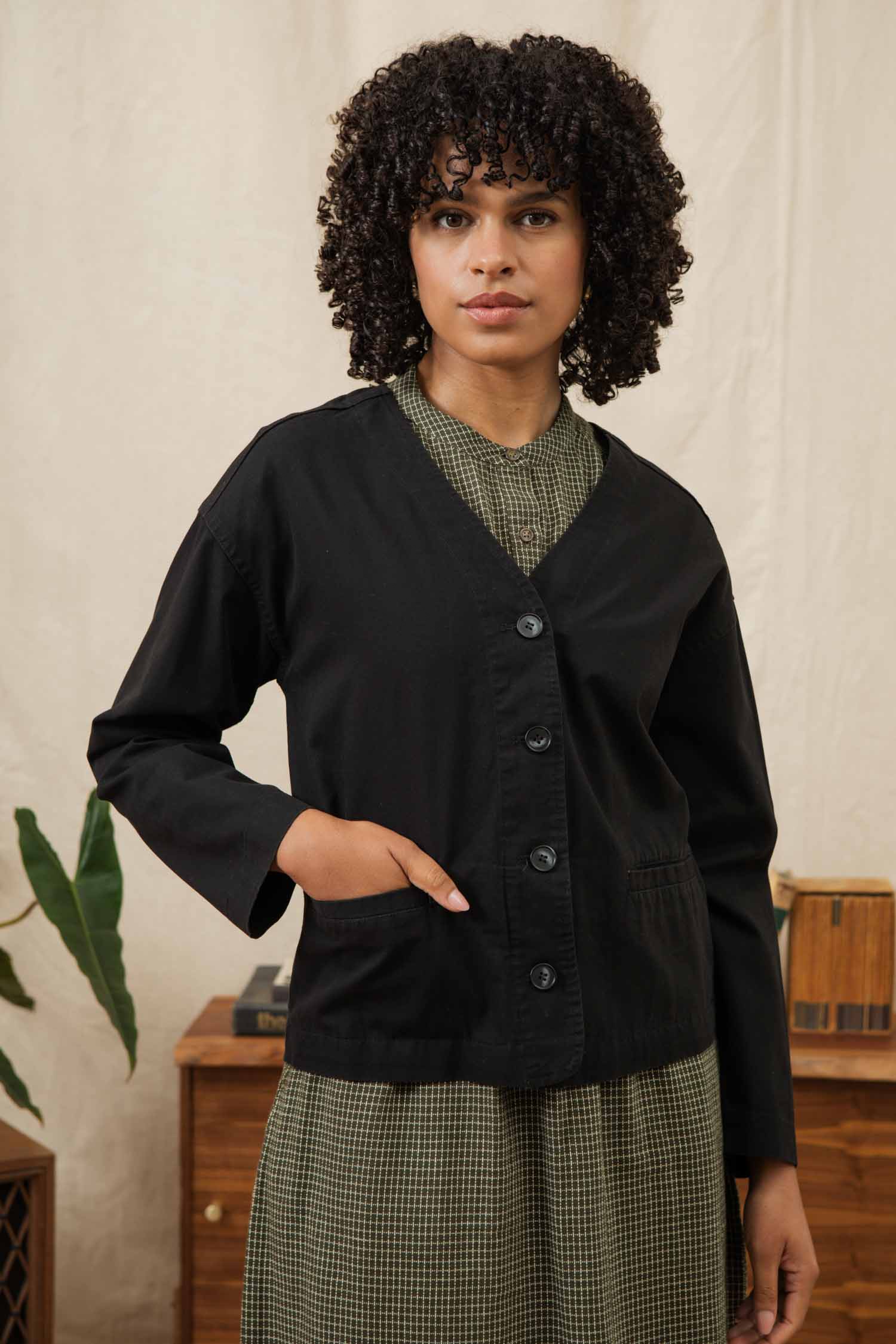 A woman with curly hair stands indoors wearing the Bridge & Burn Lee Jacket in black canvas over a green checkered dress. She has one hand in her jacket pocket and looks at the camera, with a plant and wooden furniture in the background.