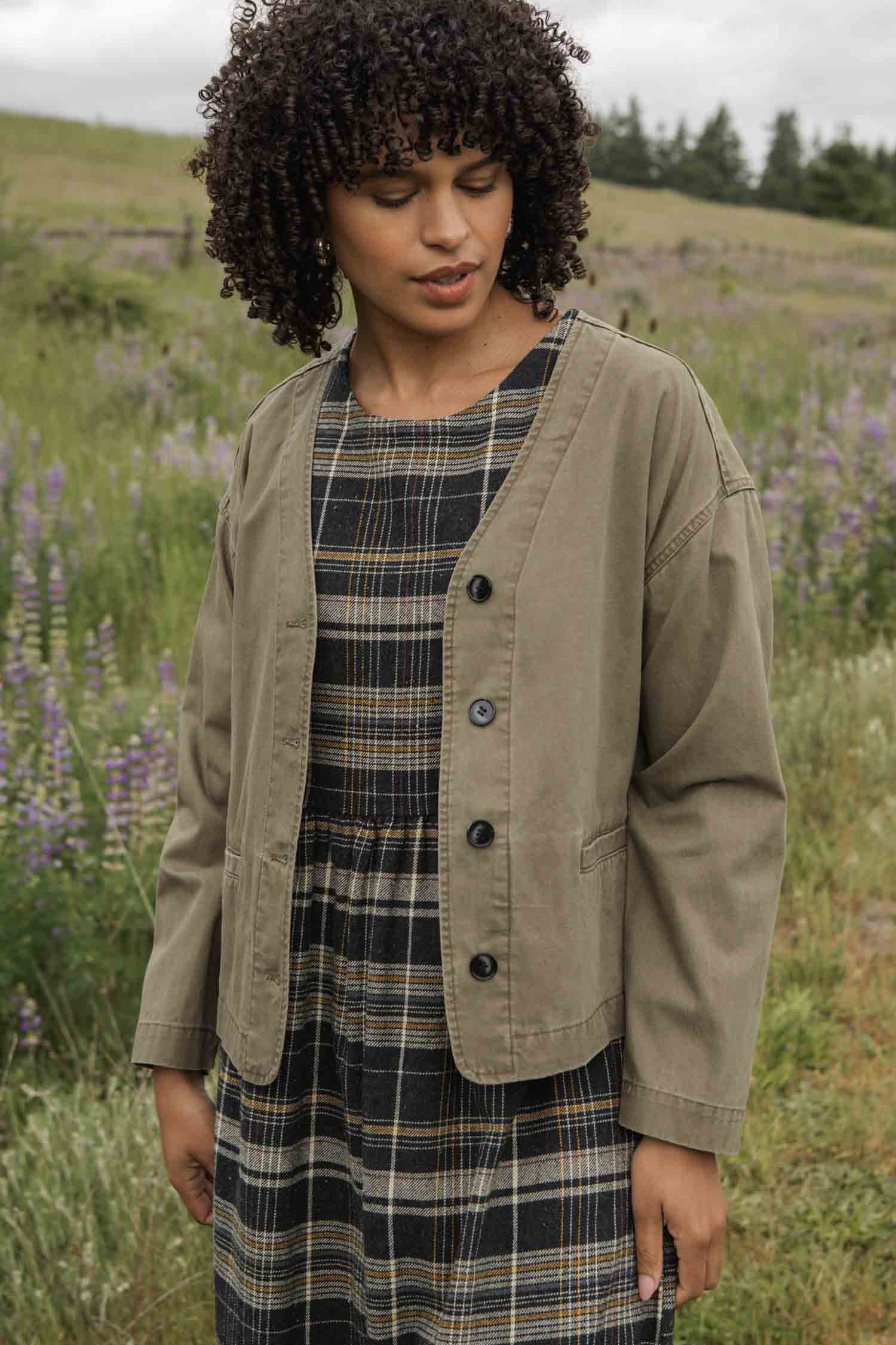 A woman with curly hair stands outdoors in a field, looking down gently as she wears the Bridge & Burn Lee Jacket in khaki green canvas over a plaid dress, surrounded by grass and purple wildflowers.