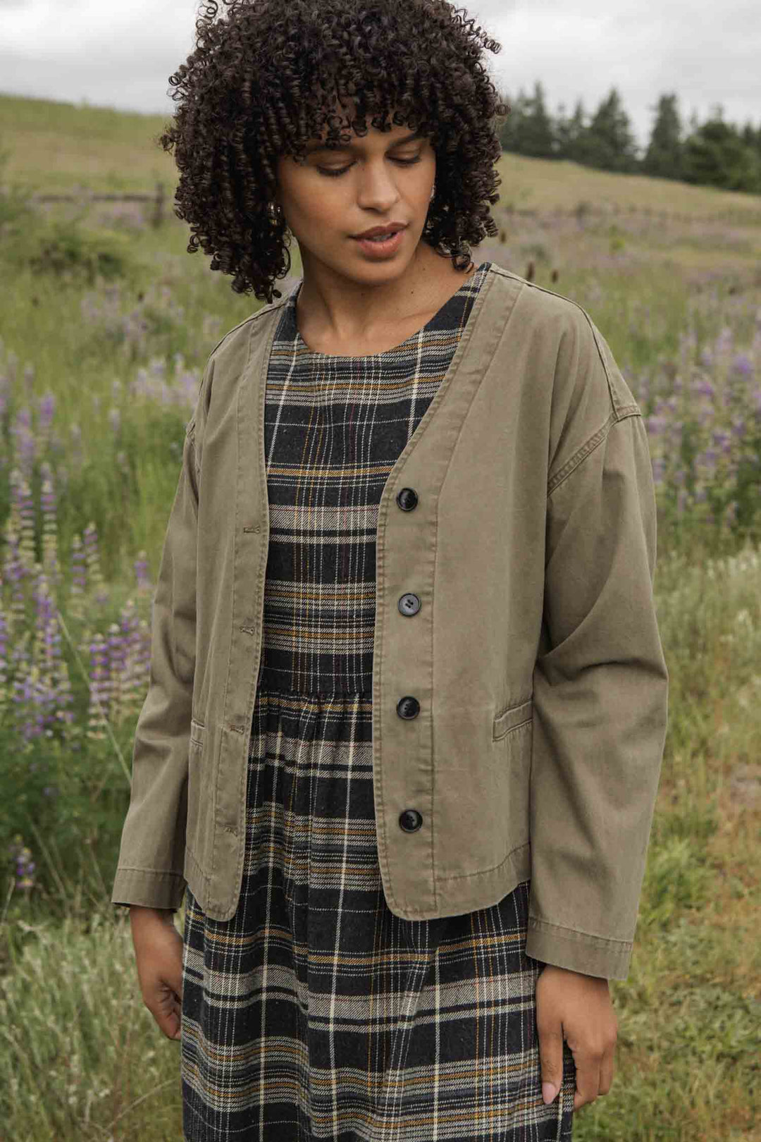 A woman with curly hair stands outdoors in a field, looking down gently as she wears the Bridge & Burn Lee Jacket in khaki green canvas over a plaid dress, surrounded by grass and purple wildflowers.