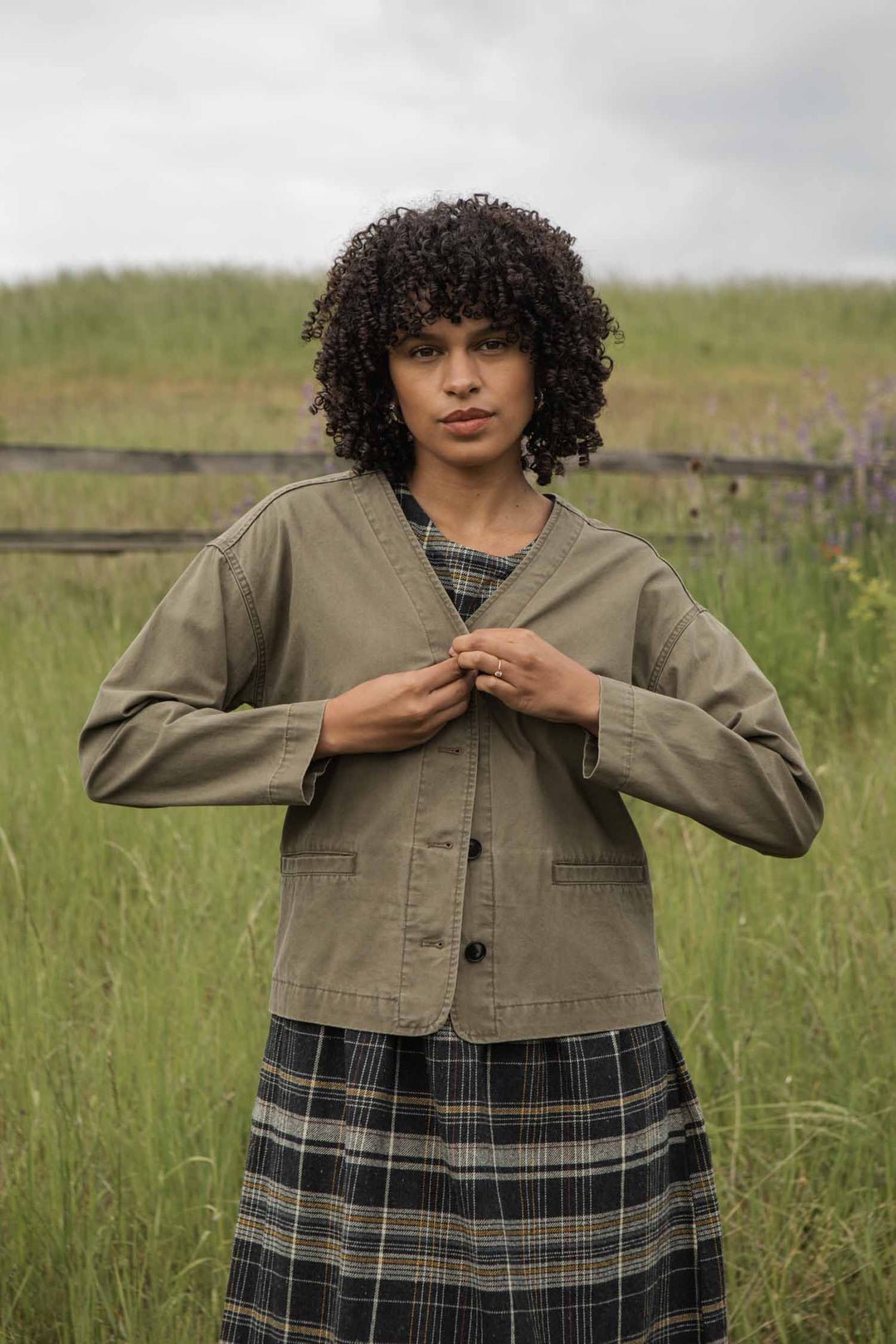 A woman with curly hair stands outdoors in a grassy field, buttoning the front of her Bridge & Burn Lee Jacket in Khaki Green Canvas. A wooden fence and cloudy sky add to the rustic backdrop.