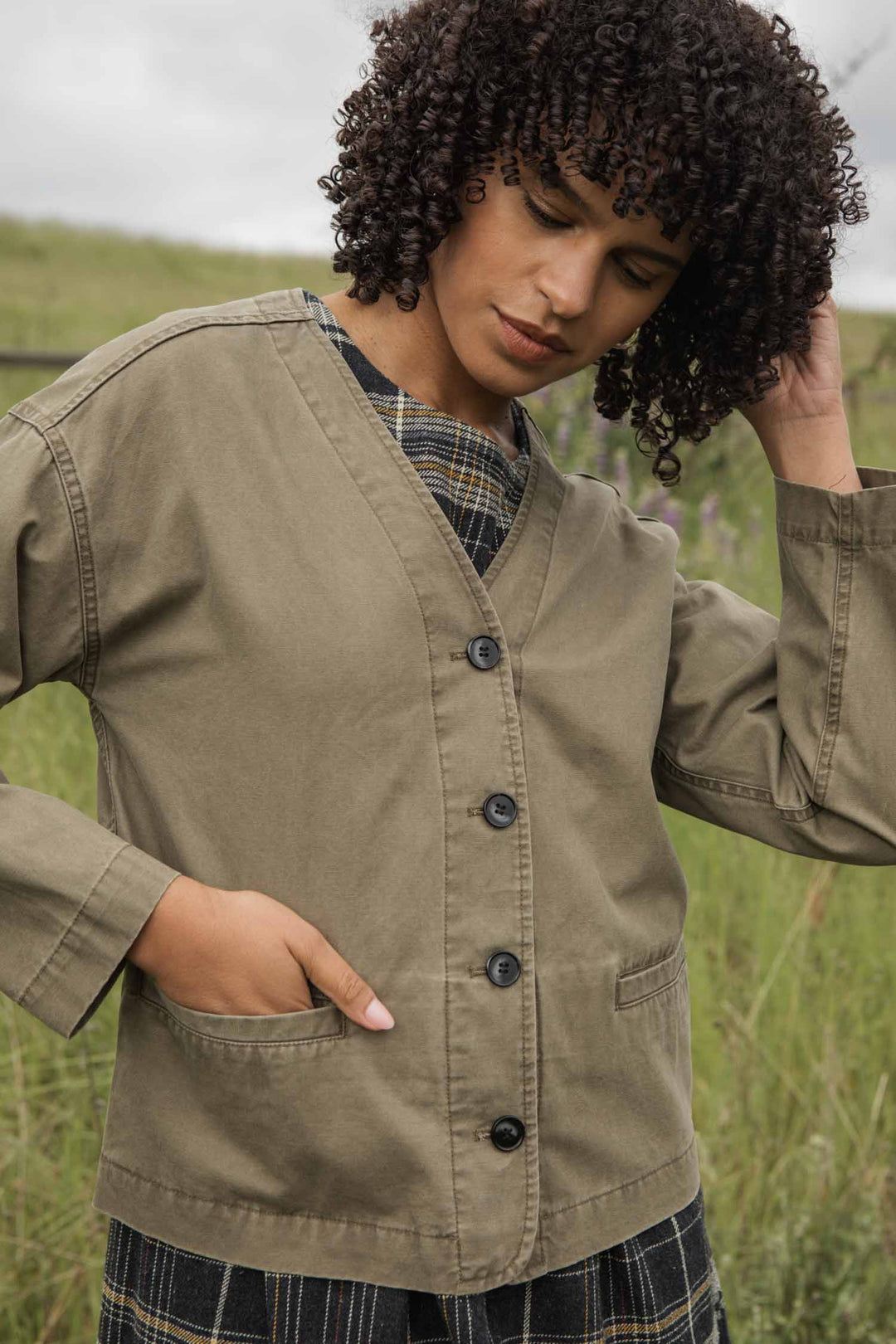 A person with curly hair wears the Bridge & Burn Lee Jacket in Khaki Green Canvas over a plaid dress, standing outdoors in a grassy field and touching their hair while looking downward.