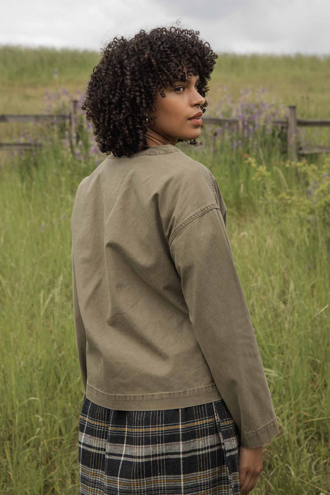 A person with curly hair stands outdoors in tall grass, wearing the Bridge & Burn Lee Jacket in Khaki Green Canvas and a plaid skirt, looking over their shoulder. A wooden fence and wildflowers are visible in the background.