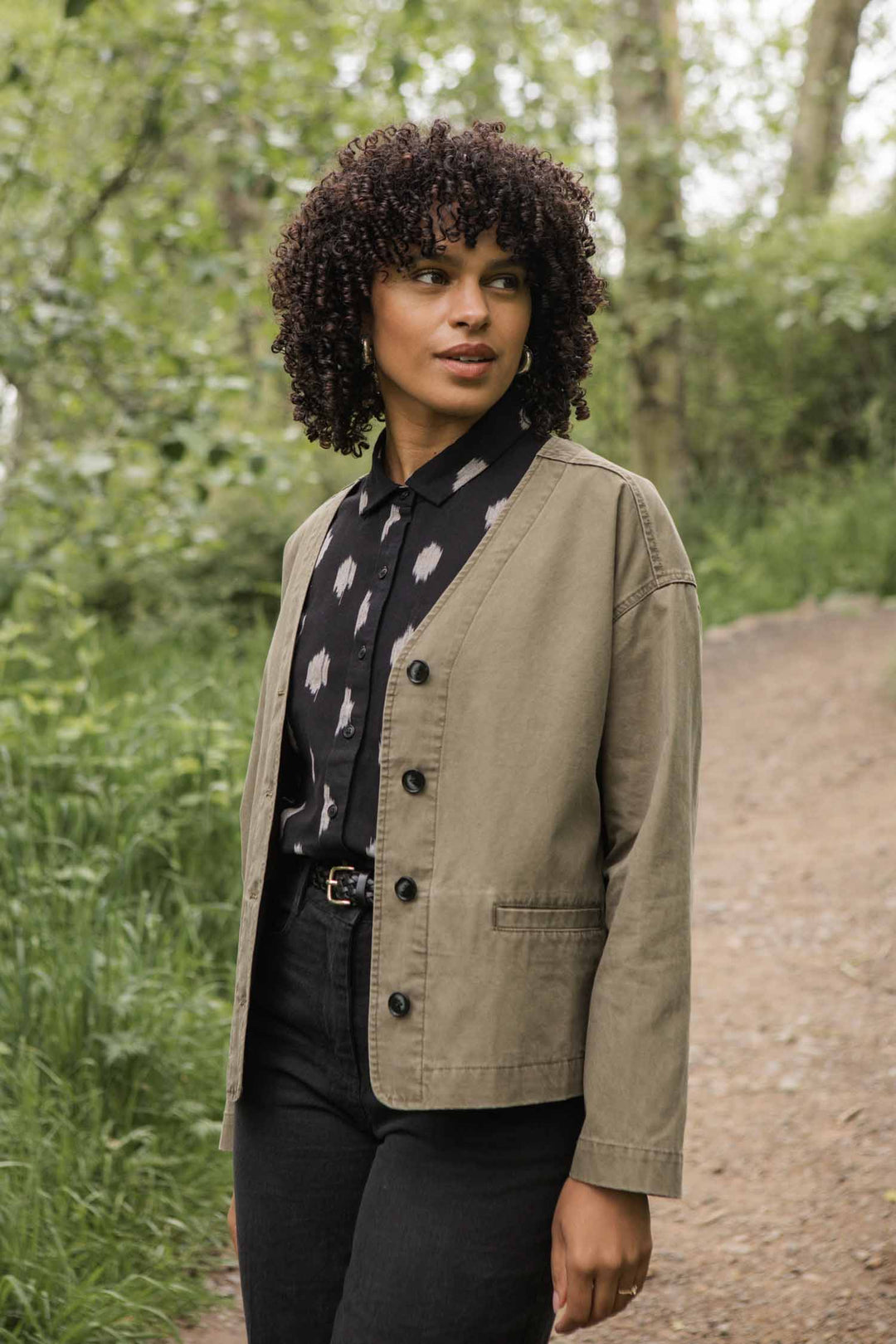 A woman with curly hair wears the Bridge & Burn Lee Jacket in Khaki Green Canvas over a black patterned shirt and black pants, standing on a dirt path surrounded by greenery and looking to the side.
