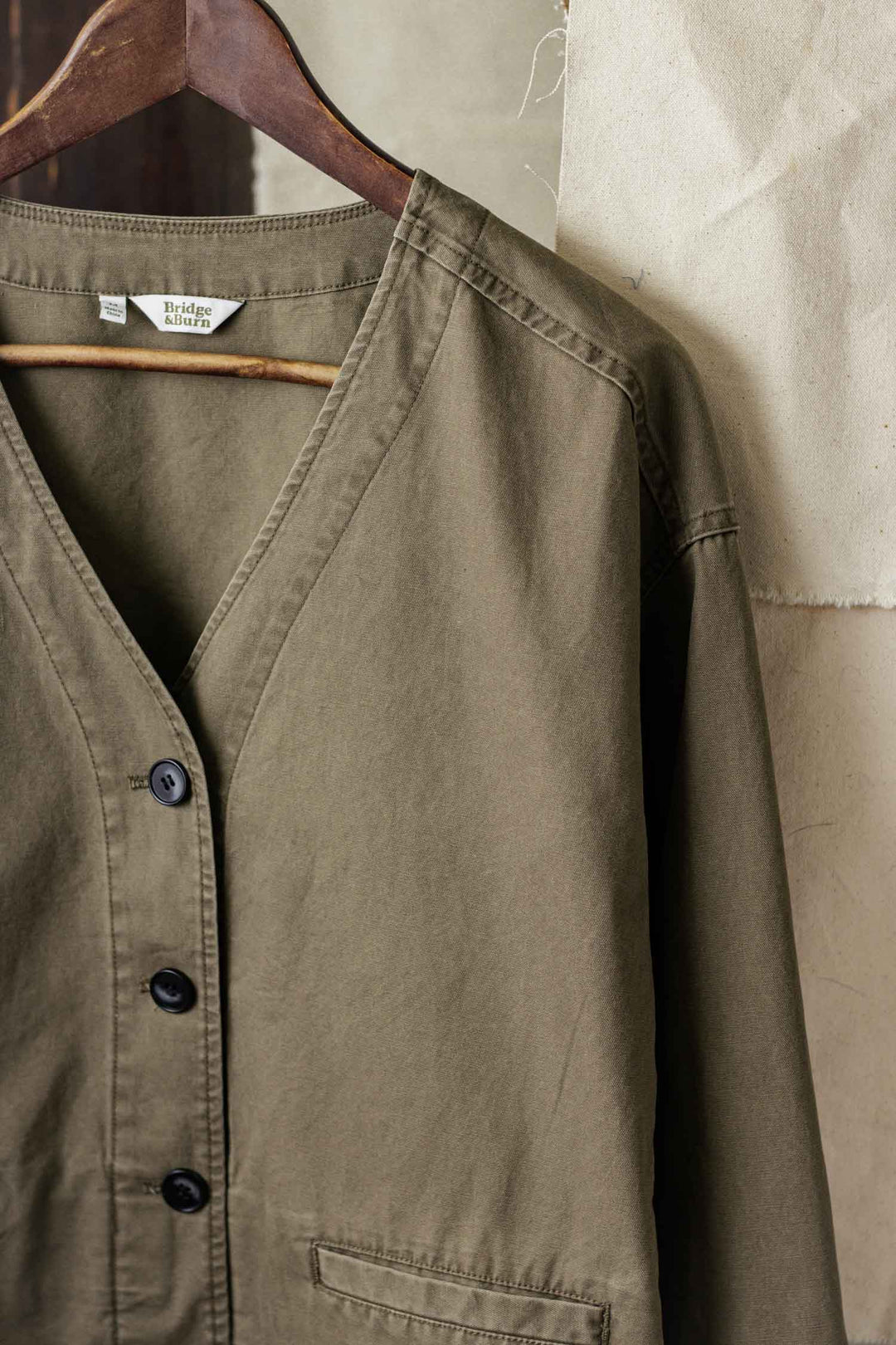 A close-up of the Bridge & Burn Lee Jacket in Khaki Green Canvas, featuring black buttons, V-neck, visible stitching, and utilitarian jacket styling. It's displayed on a wooden hanger against a light fabric background.