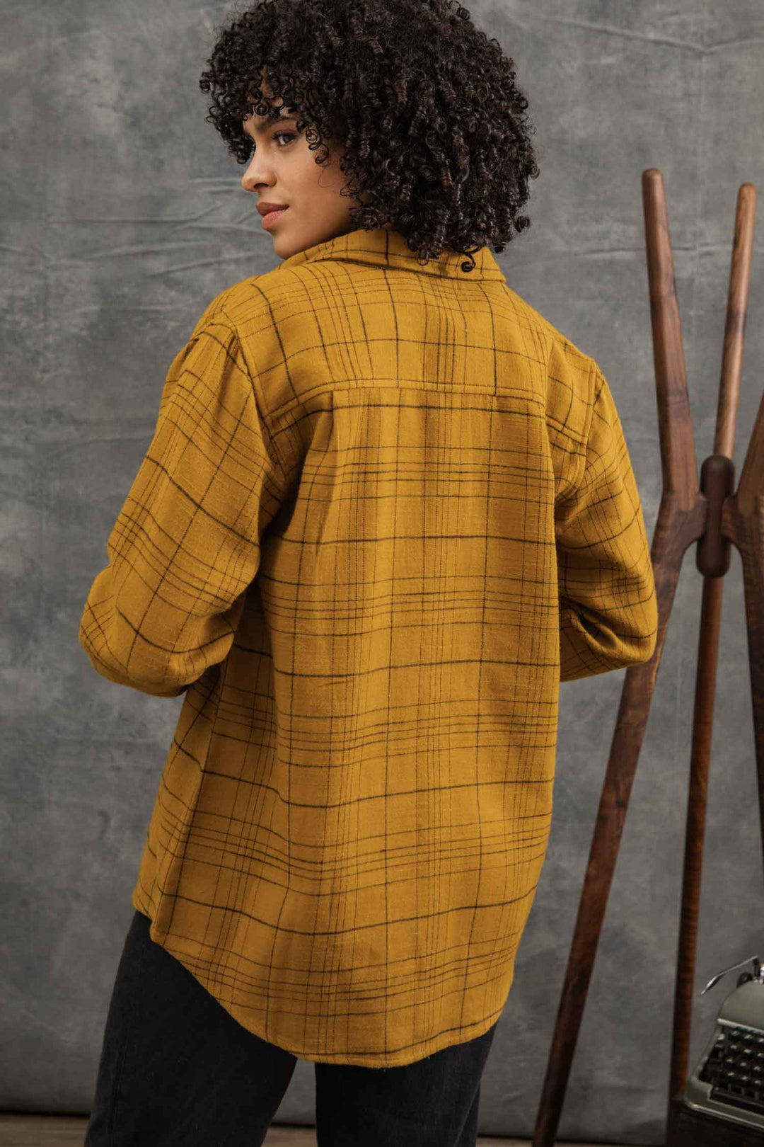 A person with curly hair models the Bridge & Burn Lenon Shirt in Golden Horizon Plaid, standing with their back to the camera and looking over their shoulder. A wooden coat rack and typewriter appear in the background.