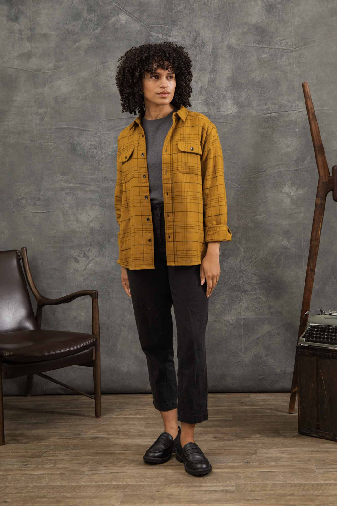 A person with curly hair stands indoors on a wooden floor near a chair, ladder, and vintage typewriter, wearing the Bridge & Burn Lenon Shirt in Golden Horizon Plaid over a gray tee, paired with black pants and shoes.
