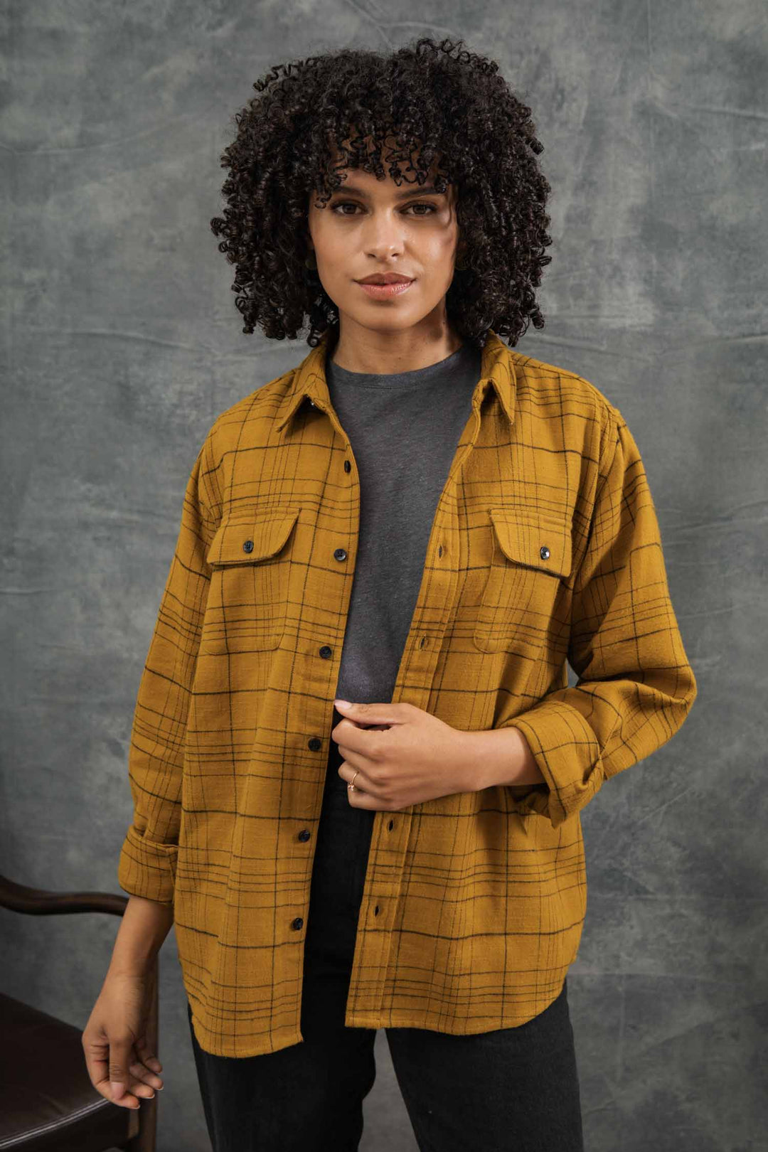 A person with curly hair wears the Bridge & Burn Lenon Shirt in Golden Horizon Plaid, a relaxed fit made from organic cotton, layered over a dark gray t-shirt, standing before a mottled gray backdrop and looking at the camera.
