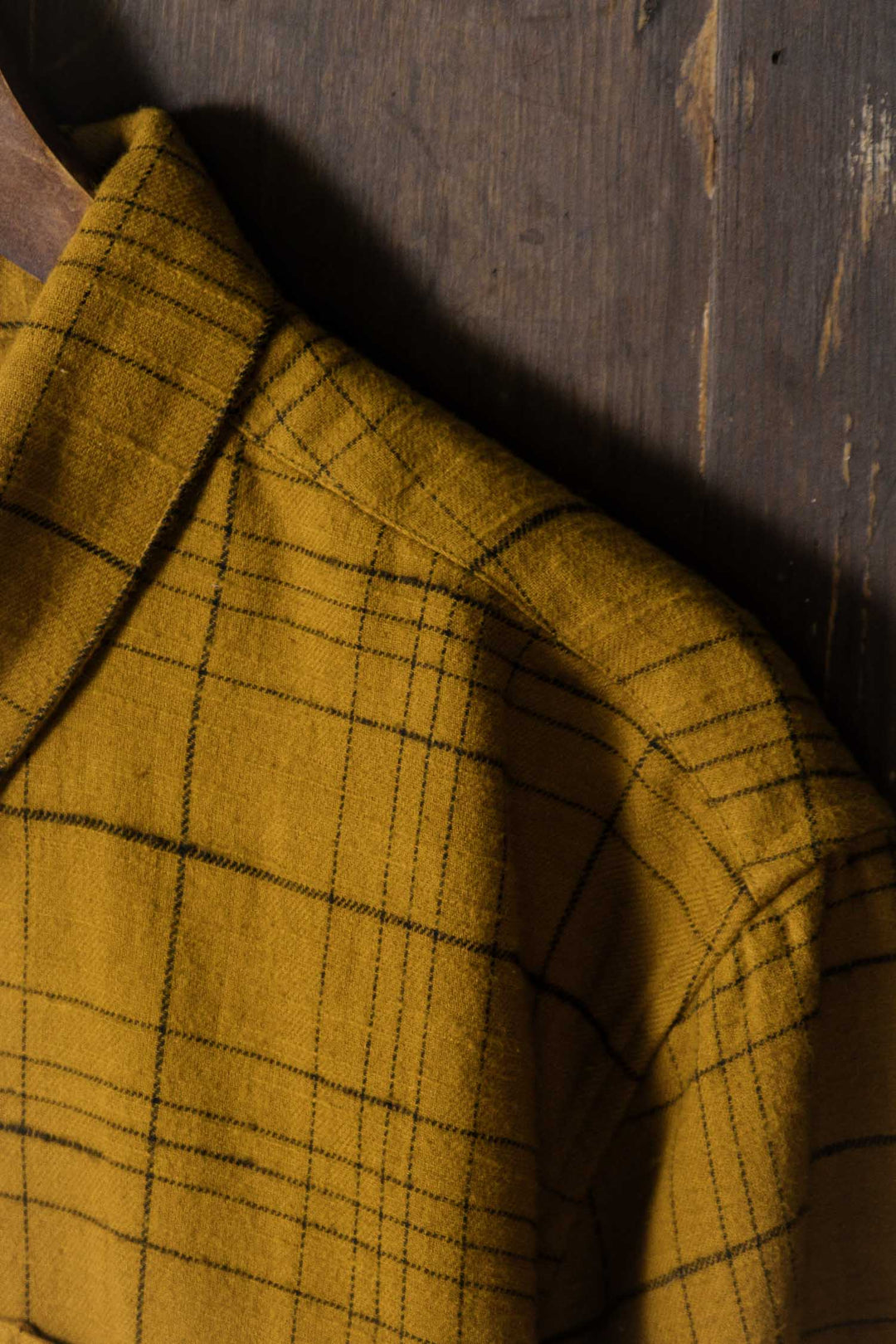 A close-up of the Lenon Shirt in Golden Horizon Plaid by Bridge & Burn, an organic cotton mustard yellow over-shirt with black plaid, displayed on a wooden hanger against a rustic wooden background.