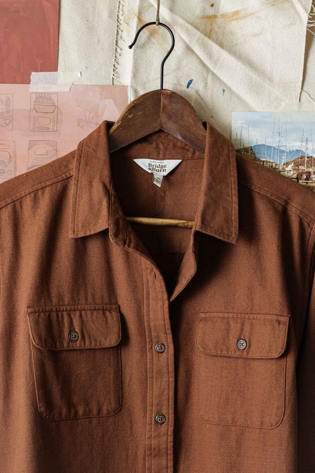 The Lenon Shirt in Terracotta Melange by Bridge & Burn, a brown organic cotton overshirt with two chest pockets, hangs on a wooden hanger against a backdrop of sketches, fabric, and boat photographs.