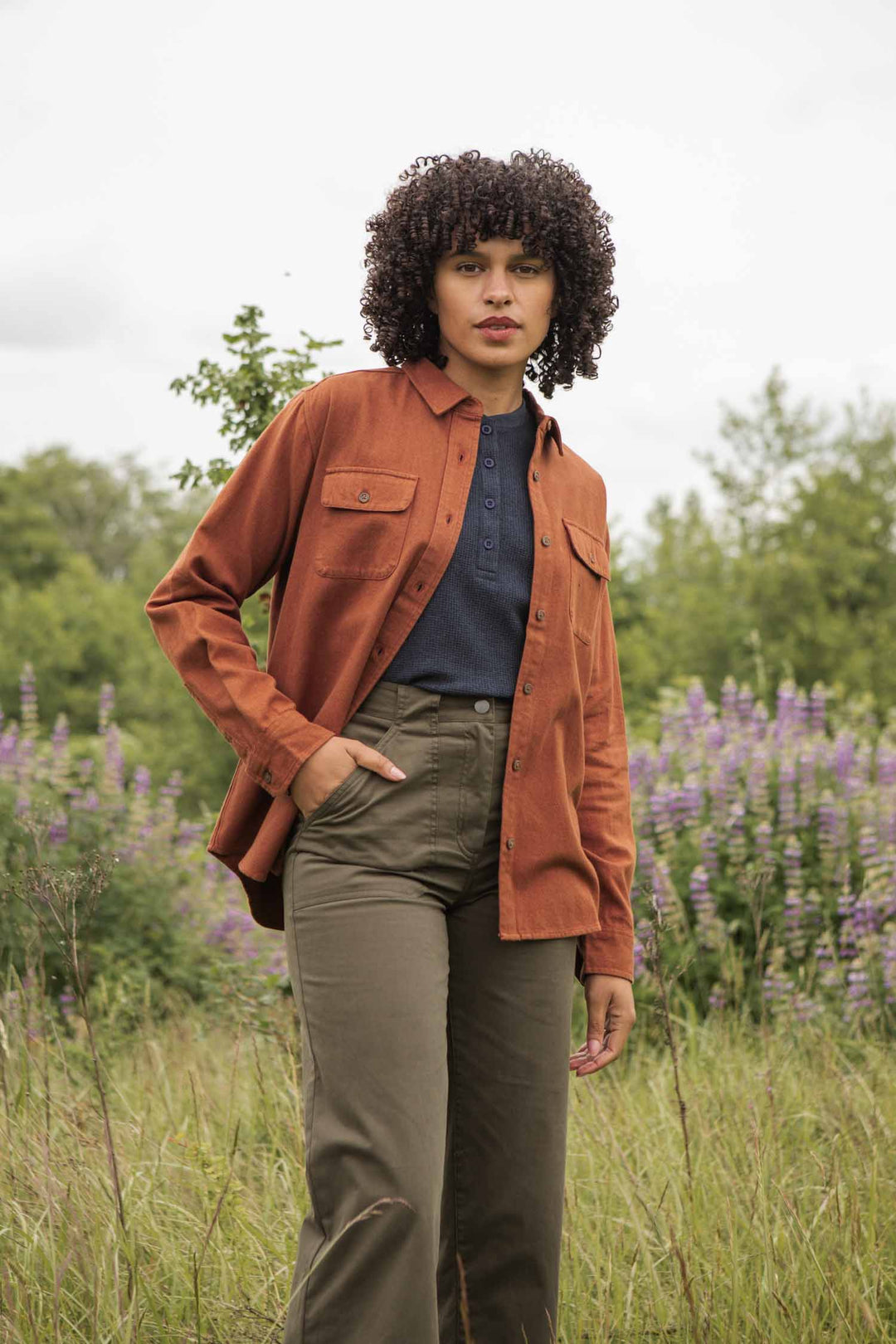 A woman with curly hair stands in a grassy field wearing the Bridge & Burn Lenon Shirt in Terracotta Melange over a navy top and olive pants, surrounded by purple flowers and green trees for a timeless backdrop.