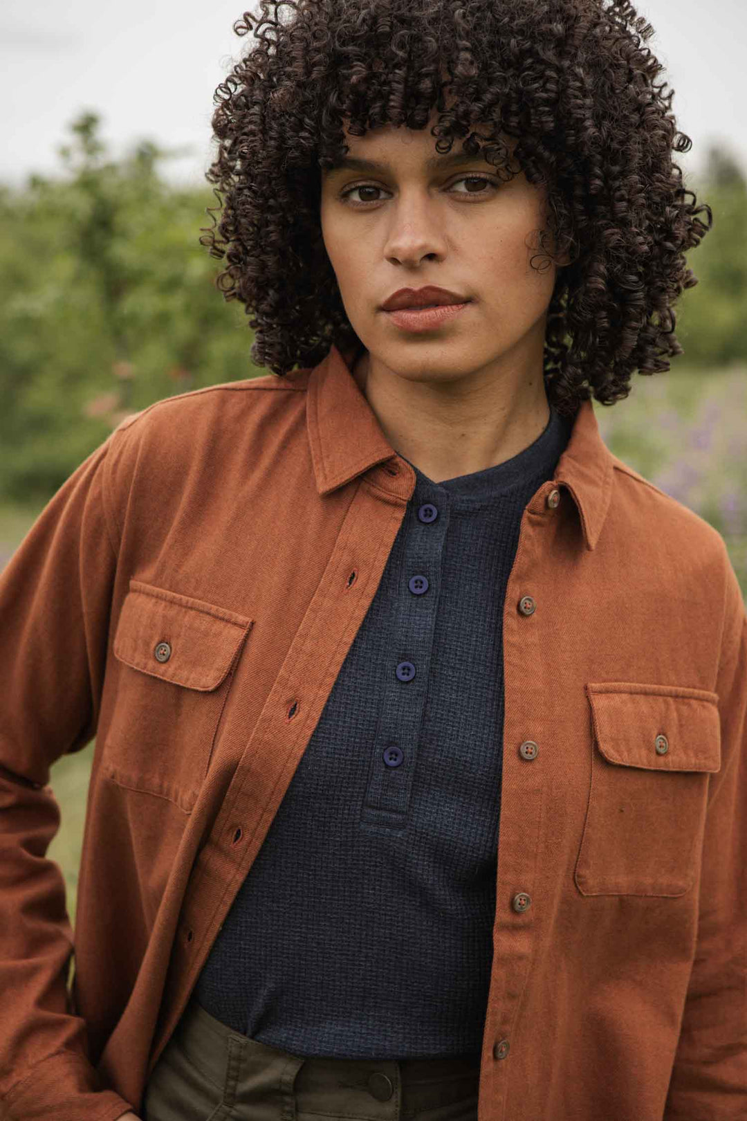 A person with curly dark hair wears the Bridge & Burn Lenon Shirt in Terracotta Melange over a navy blue top, standing outdoors with a neutral expression. Blurred greenery is visible in the background.