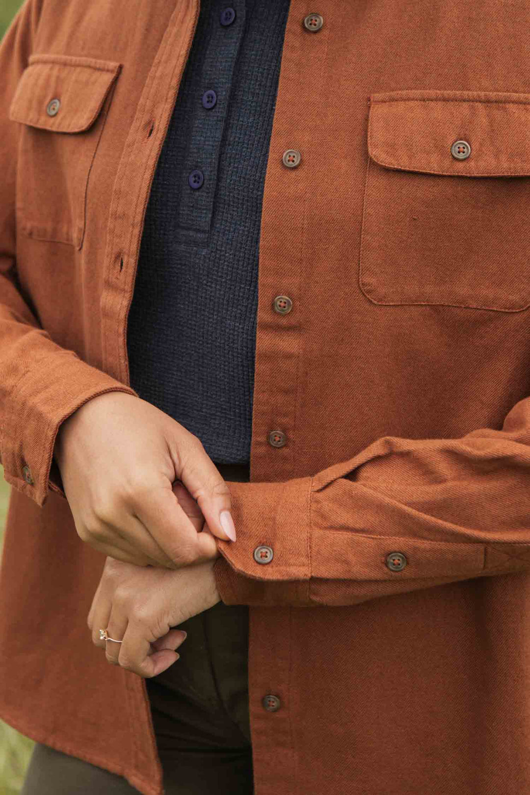 A person adjusts the cuff of the Bridge & Burn Lenon Shirt in Terracotta Melange, layered over a dark blue textured top. Only their torso and hands are visible, showcasing effortless, timeless style.